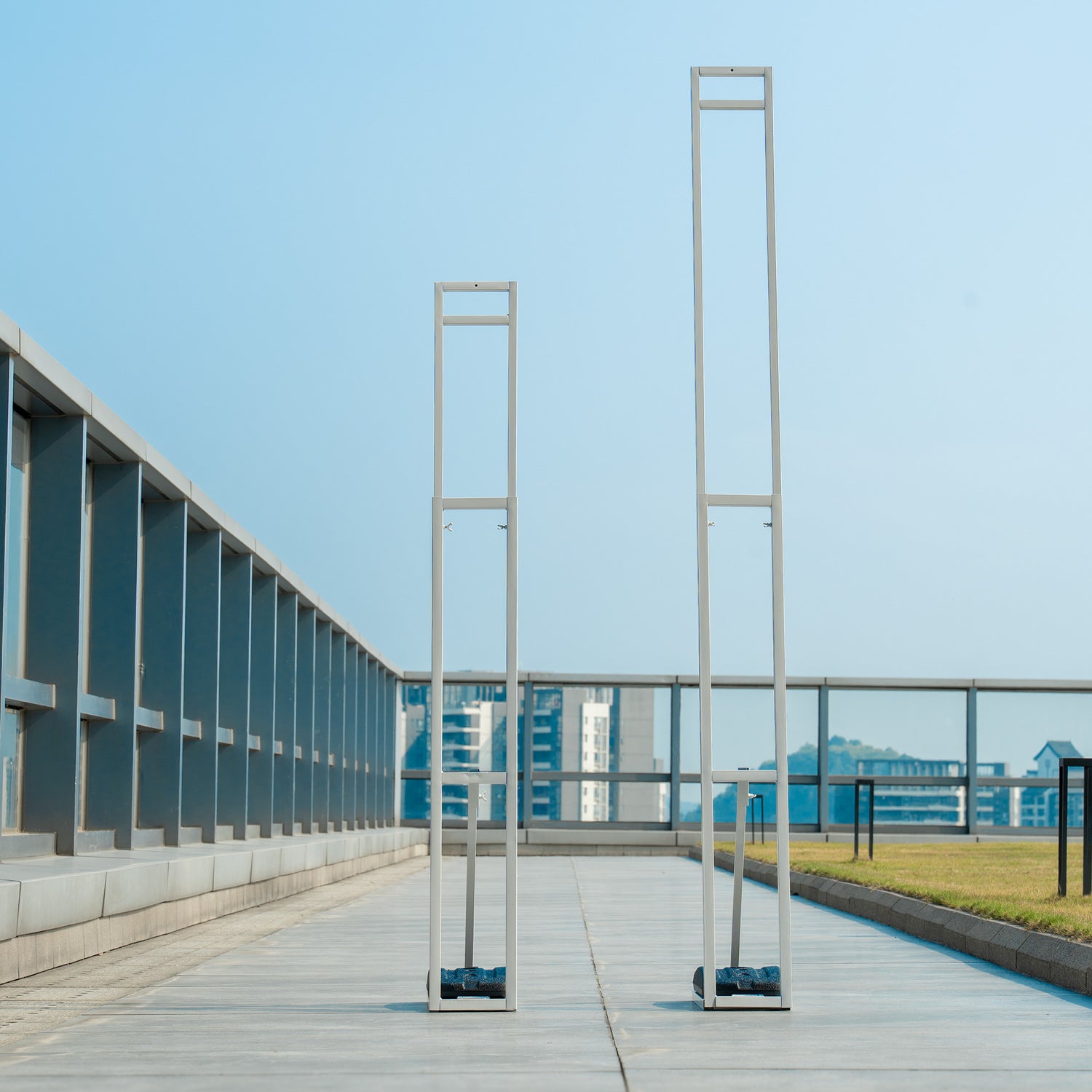 These backdrop stands are metallic structures placed on an outdoor rooftop area with a cityscape in the background. They are tall, rectangular, and minimalist in design, with a light gray finish. Each stand has horizontal bars and appears to be weighted at the base for stability, likely used to support floral or decorative backdrops.