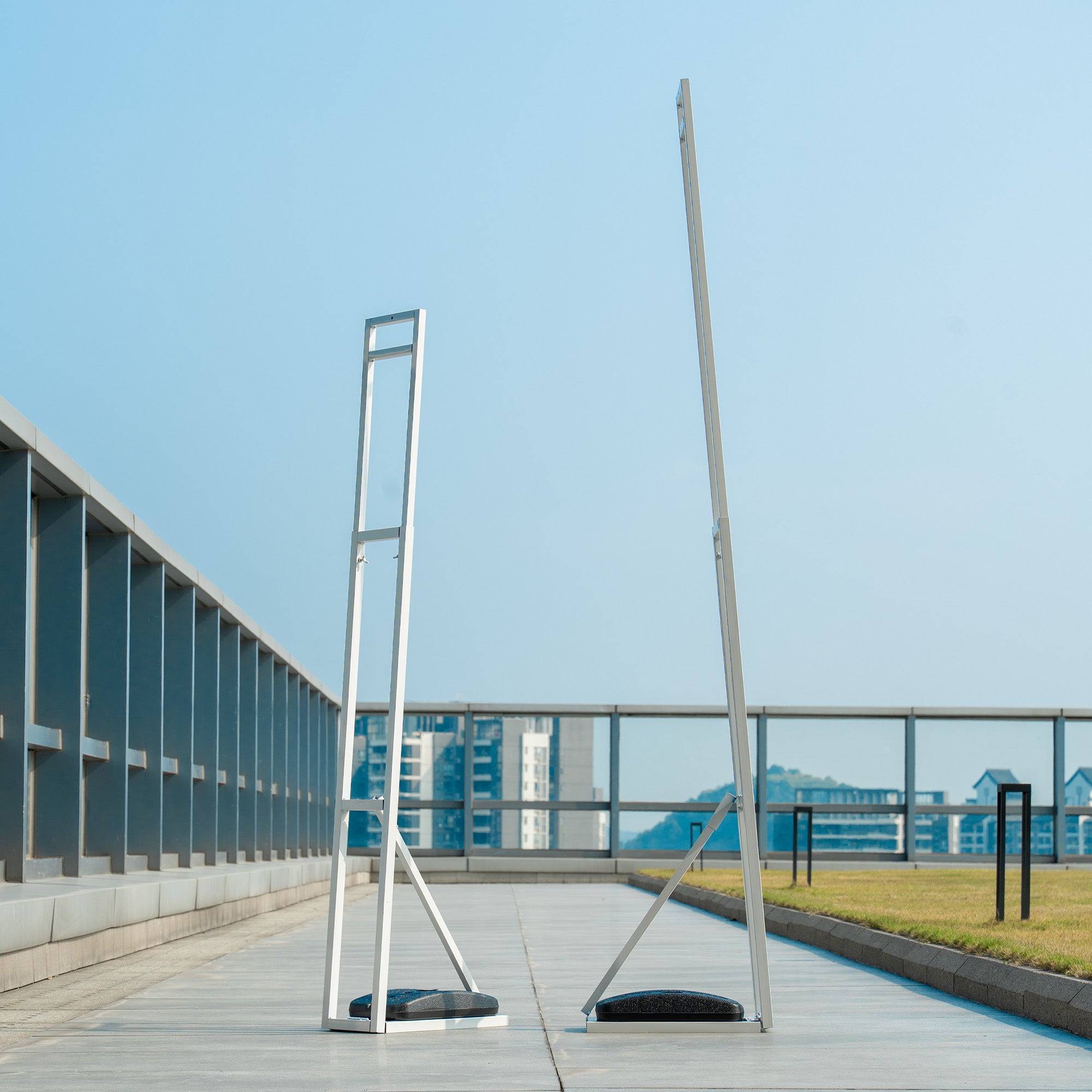 These backdrop stands are metallic structures placed on an open - air rooftop. They have a sleek, minimalist design with vertical frames and cross - supports. The stands feature weighted black bases for stability, likely intended to hold up floral or decorative backdrops for events, set against an urban cityscape background.