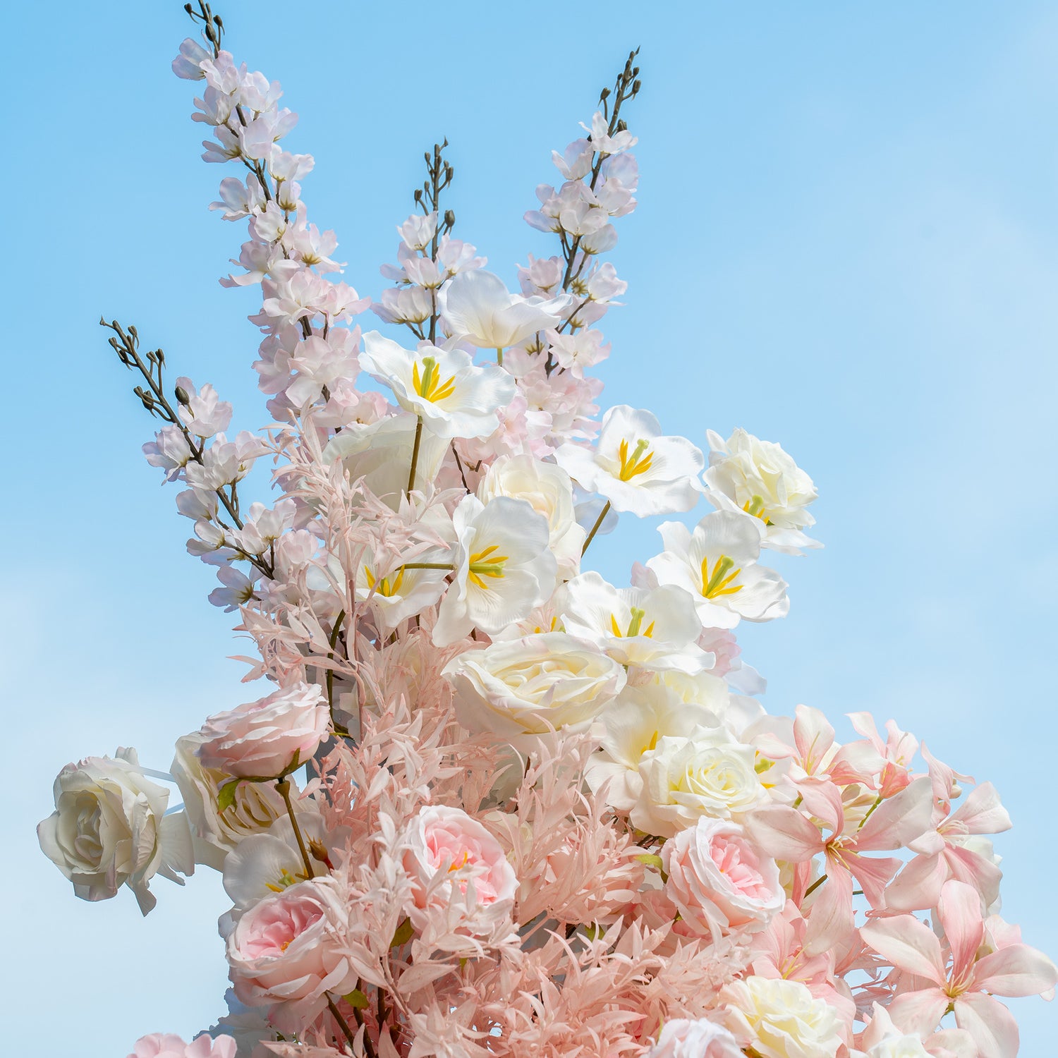 This floral arrangement features a mix of artificial flowers set against a clear blue sky. It includes white roses, pale - pink roses, white tulip - like flowers, and delicate pink blossom branches. The color palette is soft, with various shades of pink and white, creating a romantic and airy style, ideal for wedding or garden - themed events.