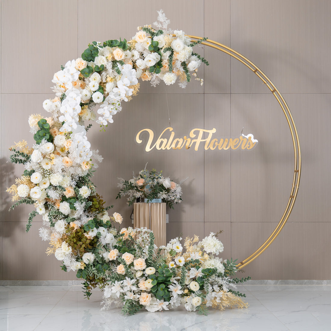 This backdrop features a golden - circular frame adorned with a lush arrangement of artificial flowers. Prominent flowers include white orchids, soft - hued roses in ivory and light peach, and white chrysanthemums. Interspersed with green eucalyptus and other foliage, the color palette is soft and elegant. The "ValarFlowers" sign adds a branded touch. The style is romantic and sophisticated, ideal for weddings or upscale events.
