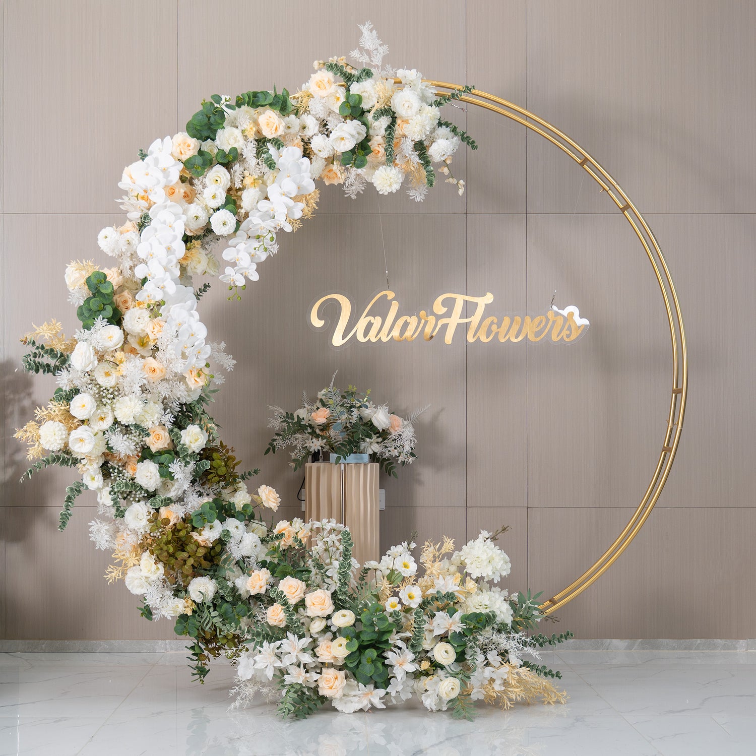 This backdrop features a golden - circular frame adorned with a lush arrangement of artificial flowers. Prominent flowers include white orchids, soft - hued roses in ivory and light peach, and white chrysanthemums. Interspersed with green eucalyptus and other foliage, the color palette is soft and elegant. The "ValarFlowers" sign adds a branded touch. The style is romantic and sophisticated, ideal for weddings or upscale events.