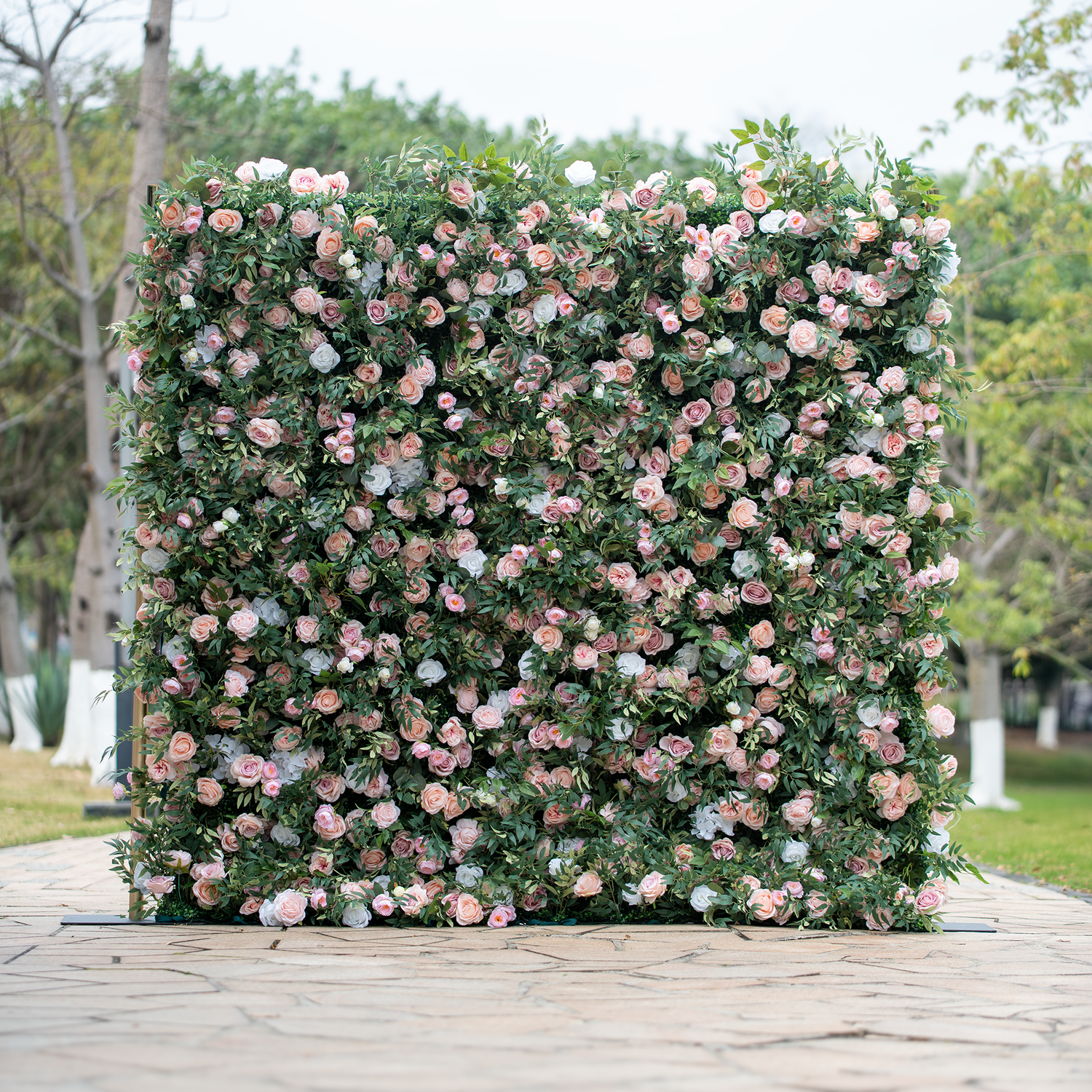 This photo showcases a large, lush floral backdrop densely adorned with an array of pink and white roses interspersed among abundant green foliage. The flowers are tightly clustered, creating a rich, textured appearance. Set outdoors on a paved area with trees in the background, it exudes a romantic and elegant ambiance, making it an ideal decorative element for weddings, garden parties, or other celebratory events.
