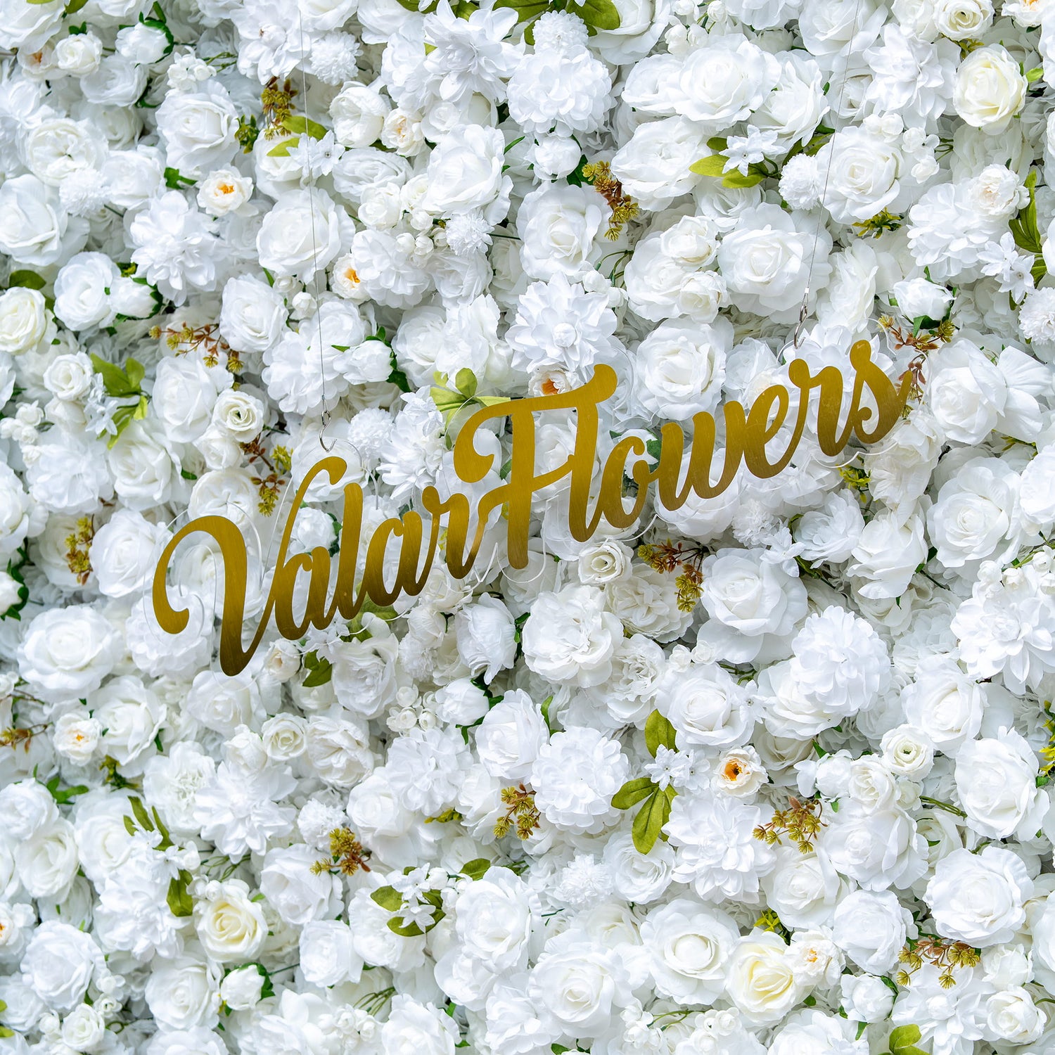 This floral backdrop is densely covered with artificial white flowers, mainly roses and some small - bud blooms, interspersed with green leaves. The all - white color scheme, enhanced by the green accents, creates a clean and elegant look. The golden “ValarFlowers” sign adds a touch of sophistication, making the overall style refined and suitable for formal events.