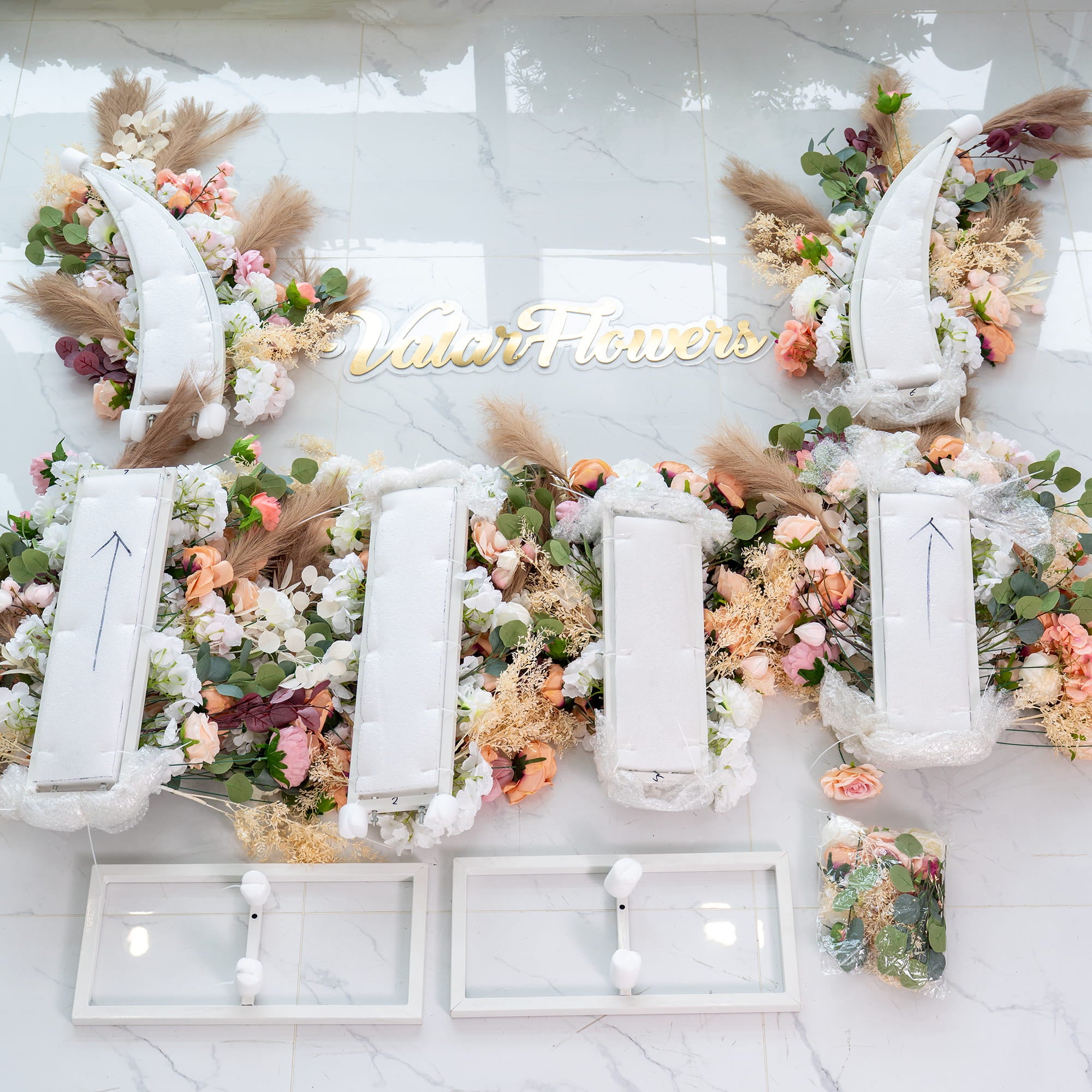 This setup features white, modular floral - arrangement structures adorned with an array of artificial flowers in soft pastel tones, including pink, peach, and white. Pampas grass and eucalyptus leaves add texture. The “Valar Flowers” branding is prominently displayed, suggesting a wedding or event decor.