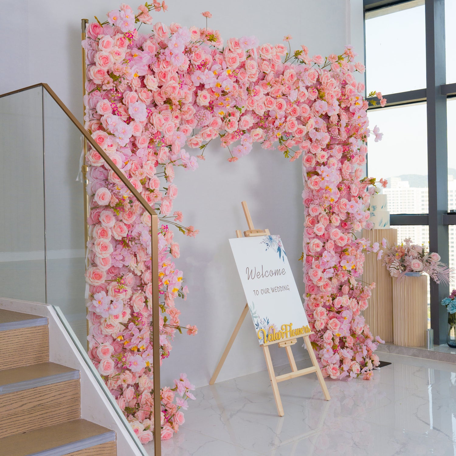 This backdrop is a lush, floral - filled archway crafted from an abundance of artificial pink roses and other assorted blossoms in soft pastel shades. It exudes a romantic and inviting atmosphere. 
In front of it stands an easel with a sign that reads "Welcome to our wedding," indicating its use for nuptial celebrations. The golden - hued "Valar Flowers" adds a touch of elegance. Ideal for creating a charming and memorable entrance at weddings.