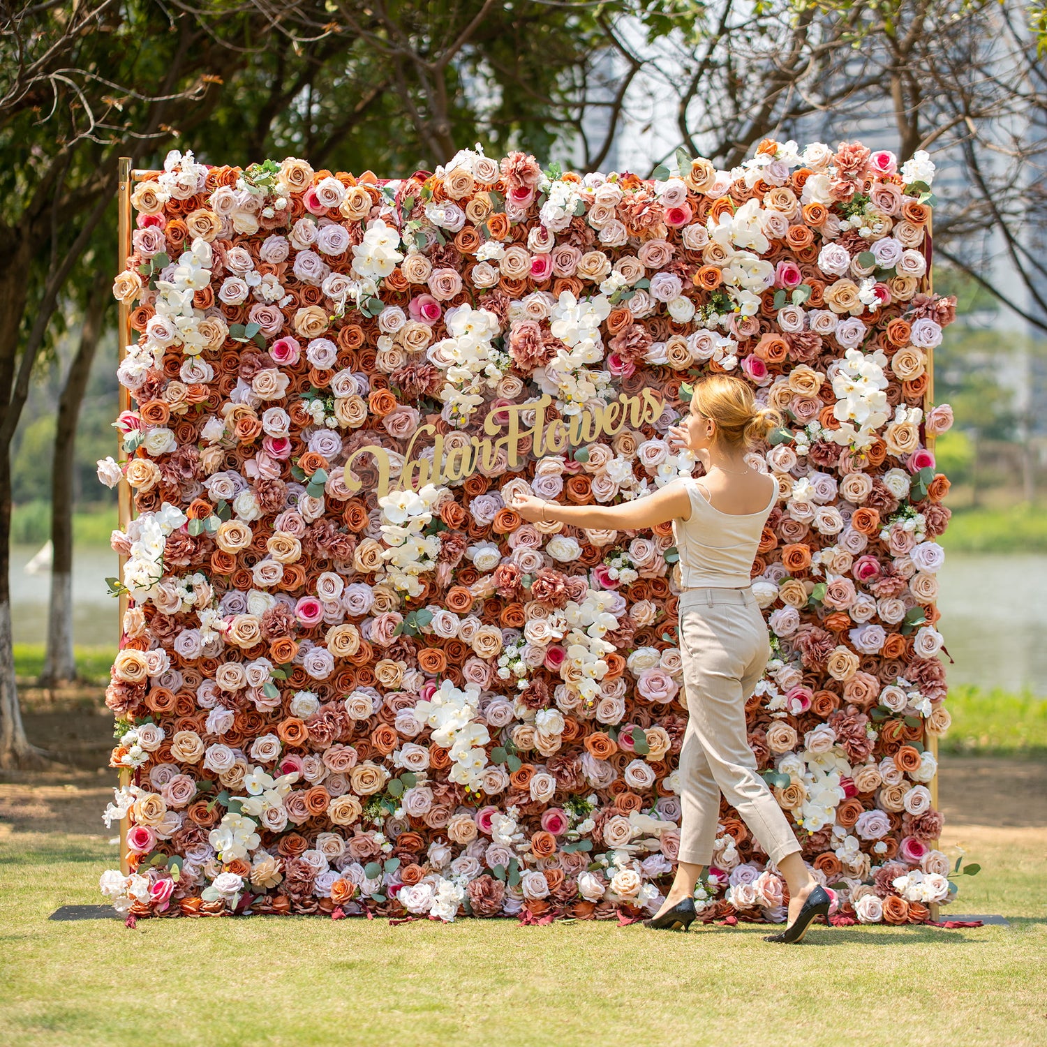 A woman with blonde hair, dressed in a light - colored sleeveless top and beige pants, stands beside a lush floral backdrop. The backdrop is filled with roses and orchids in earthy tones, with "Valar Flowers" signage, set against a park - like outdoor scene.