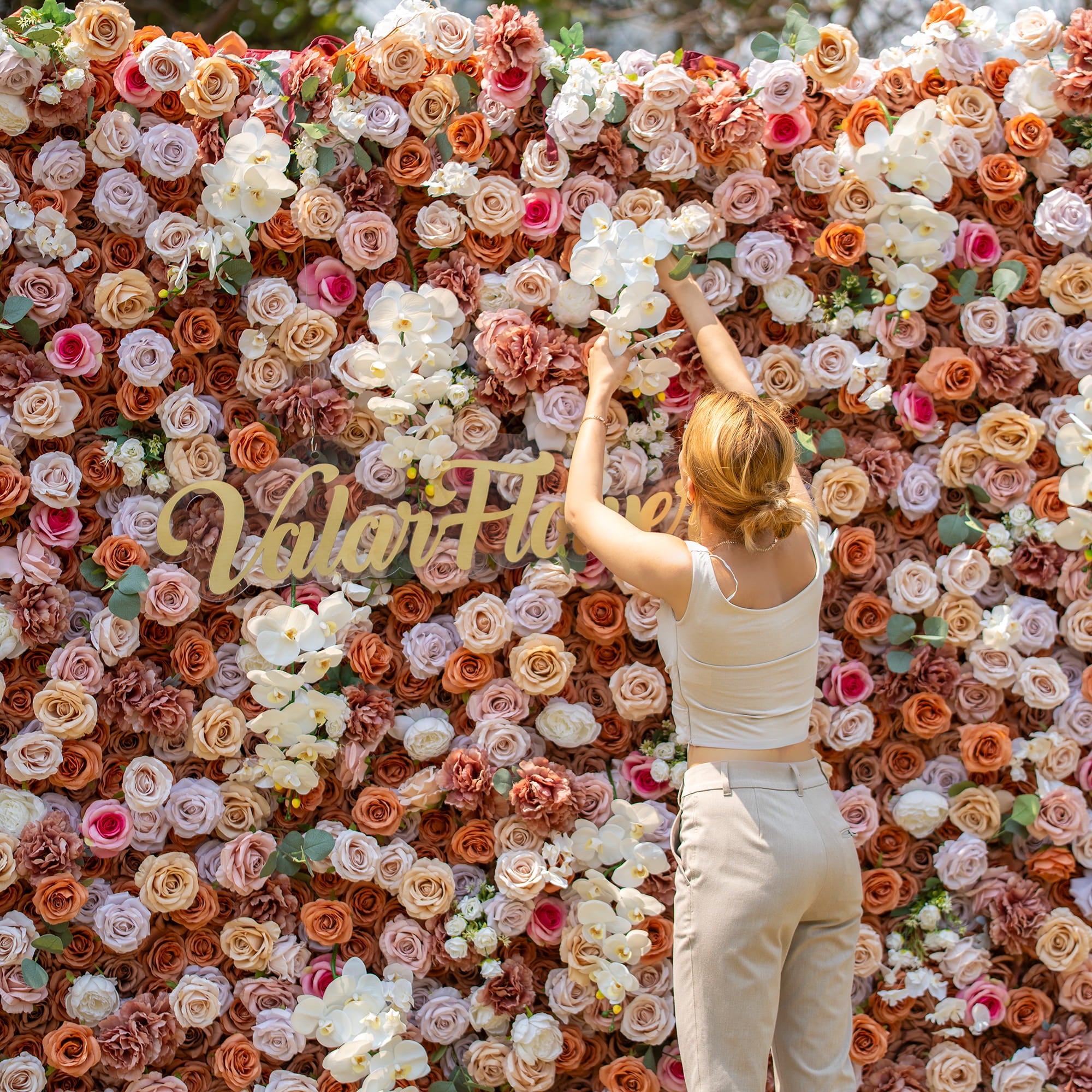A woman with blonde hair, dressed in a sleeveless top and beige pants, is arranging flowers on a lush floral backdrop. The backdrop is filled with roses and orchids in earthy tones, and features the text "Valar Flowers" in elegant gold lettering.