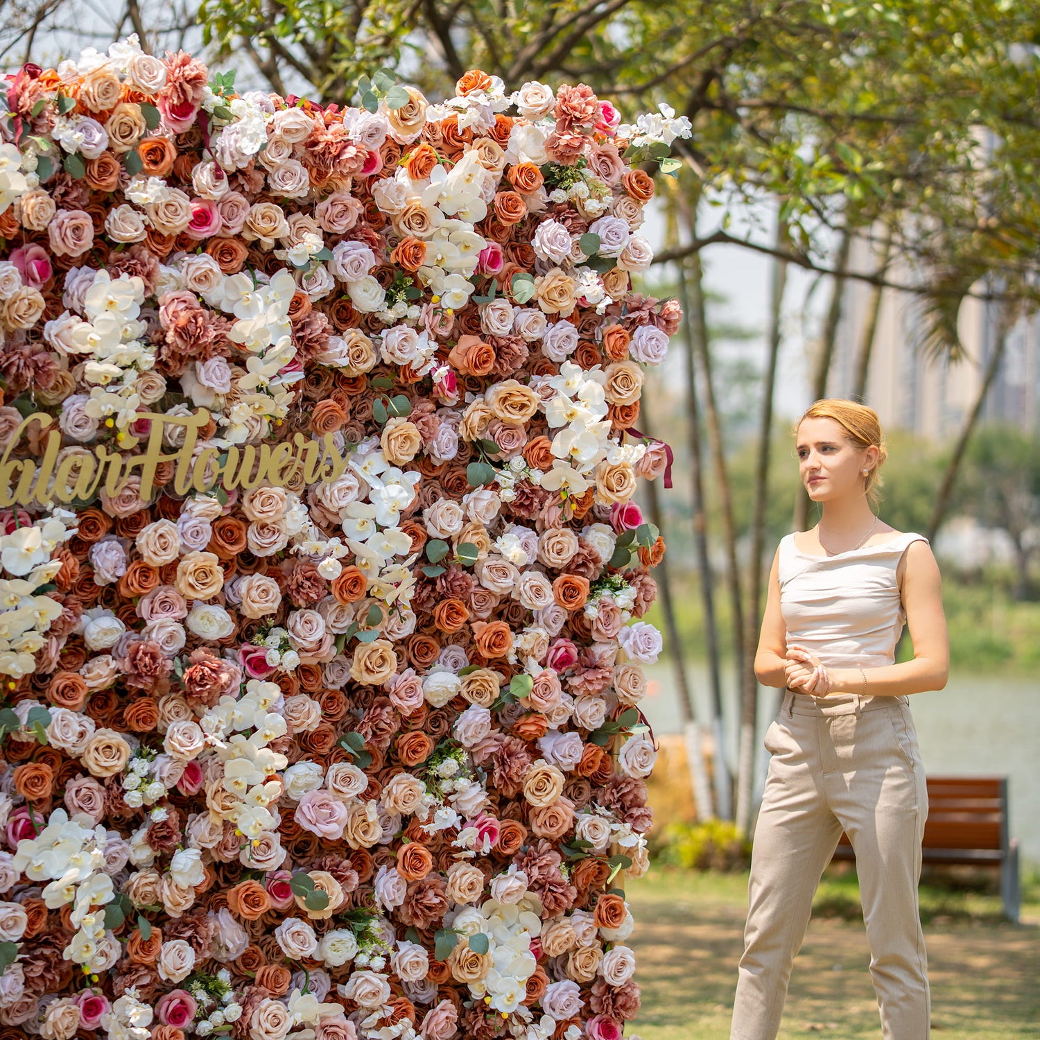 A woman with blonde hair, wearing a sleeveless top and beige pants, stands beside a lush floral backdrop. The backdrop is densely packed with roses and orchids in earthy tones, with "Valar Flowers" signage, set in an outdoor park - like environment.