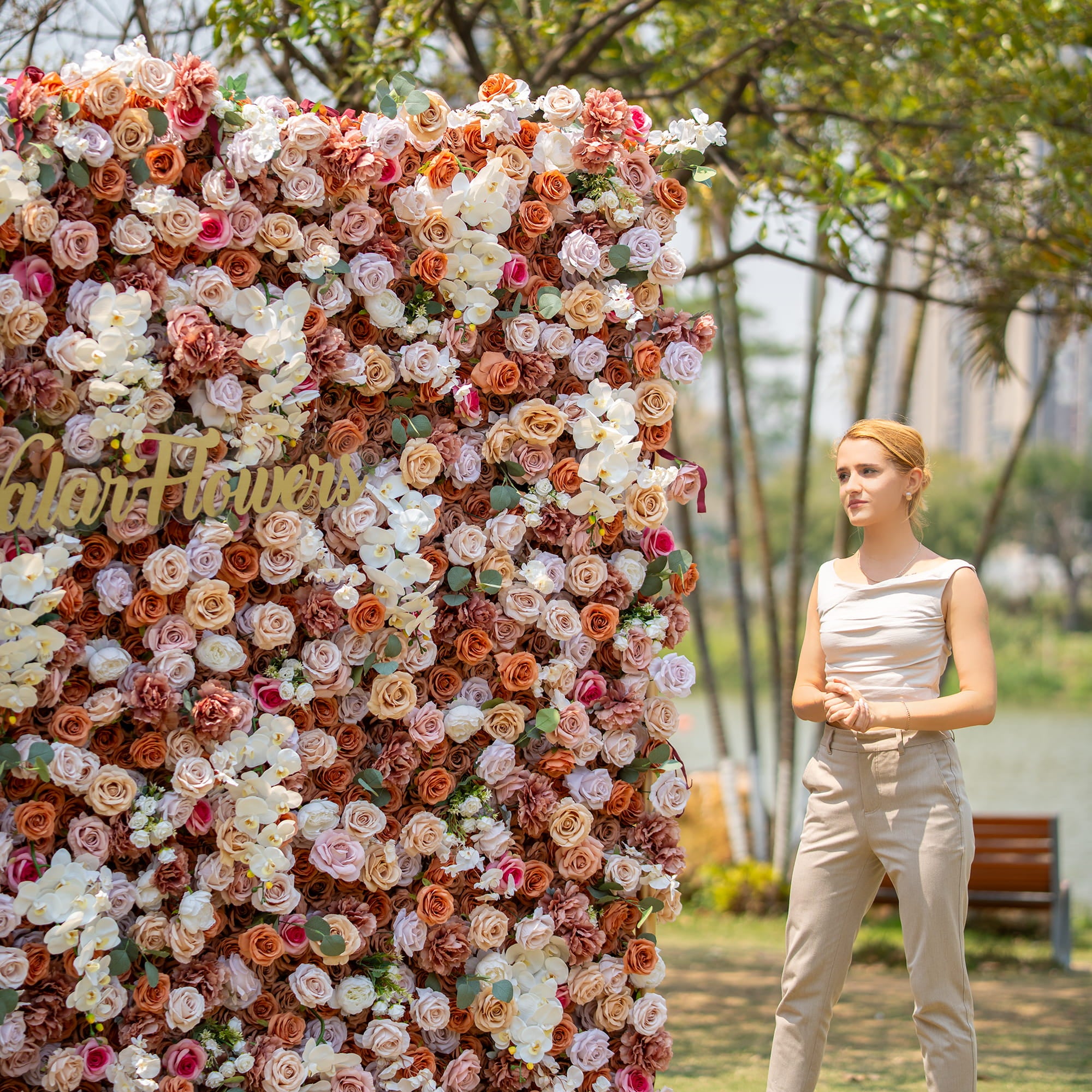 A woman with blonde hair, wearing a sleeveless top and beige pants, stands beside a lush floral backdrop. The backdrop is densely packed with roses and orchids in earthy tones, with "Valar Flowers" signage, set in an outdoor park - like environment.