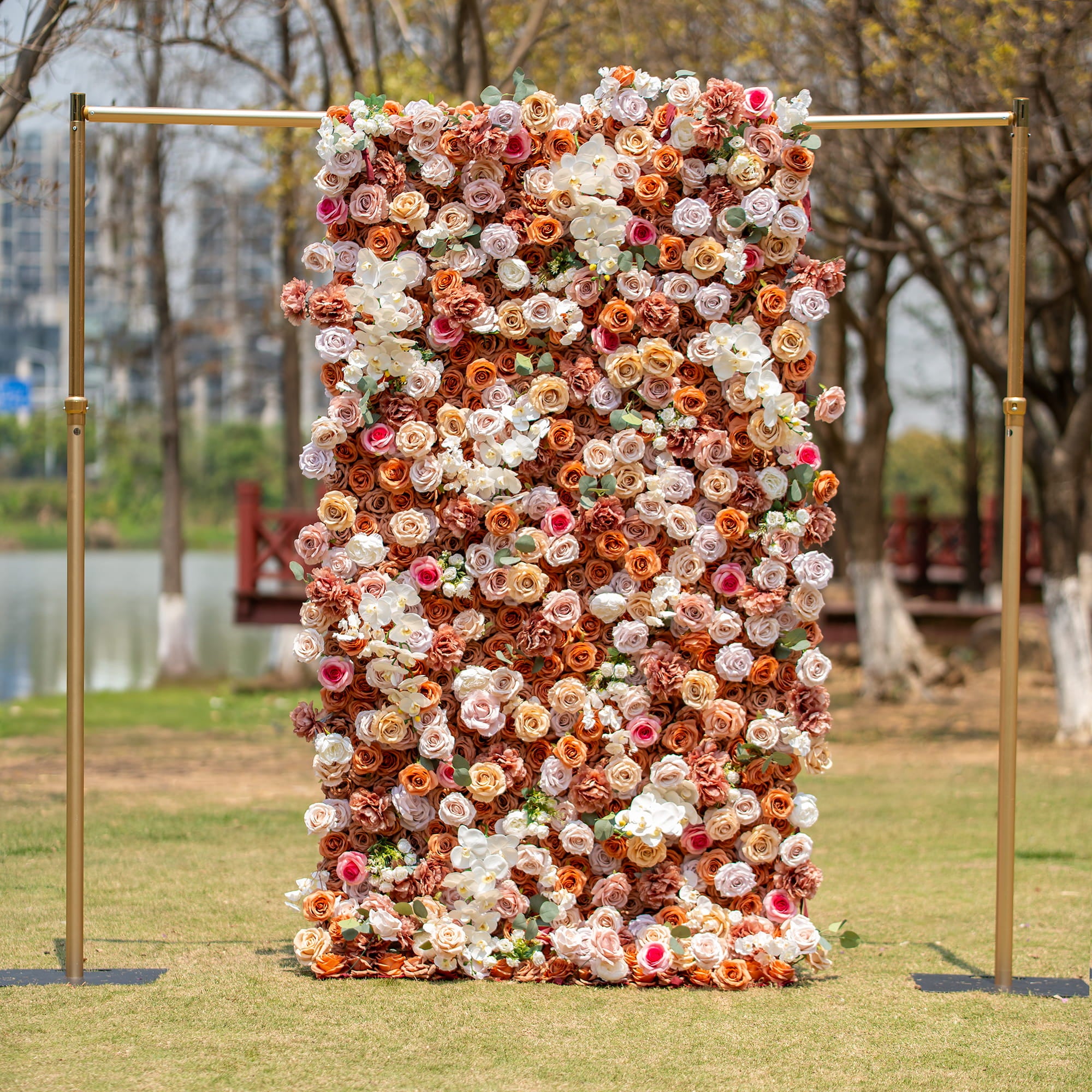The photo shows a 1.2m x 2.4m floral backdrop densely adorned with roses and orchids in earthy tones like beige, brown, and white. Set outdoors by a lake with trees and buildings in the background, it exudes elegance, ideal for events.