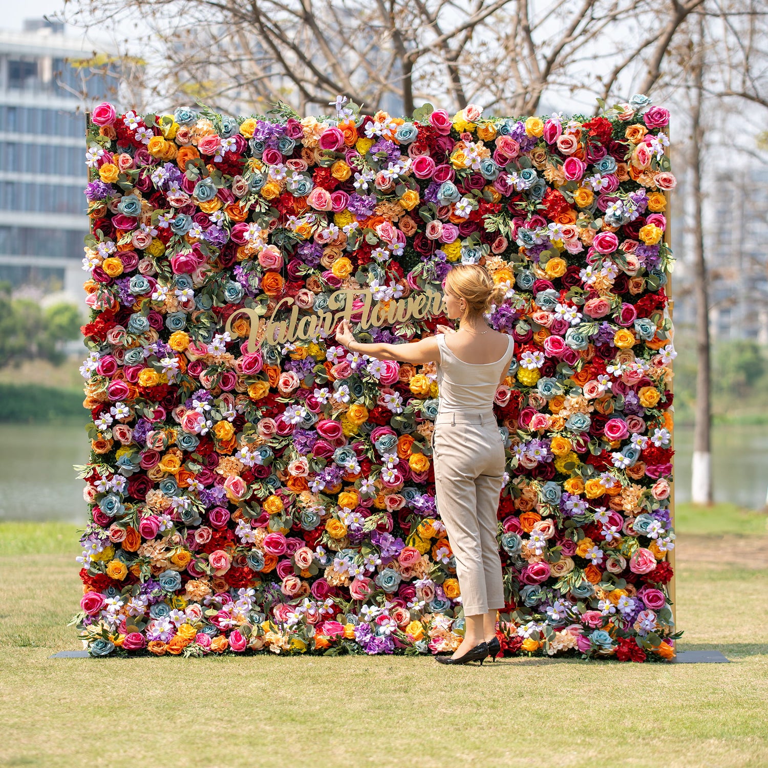 A woman in a light - colored outfit adjusts the golden “Valar Flowers” sign on a vibrant floral backdrop. The backdrop is a riot of multicolored roses and assorted blooms, set against an outdoor park - like scene.