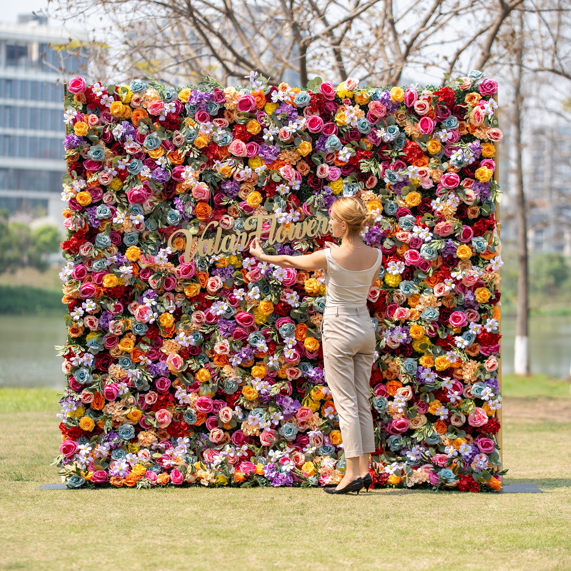 A woman in a light - colored outfit adjusts the golden “Valar Flowers” sign on a vibrant floral backdrop. The backdrop is a riot of multicolored roses and assorted blooms, set against an outdoor park - like scene.