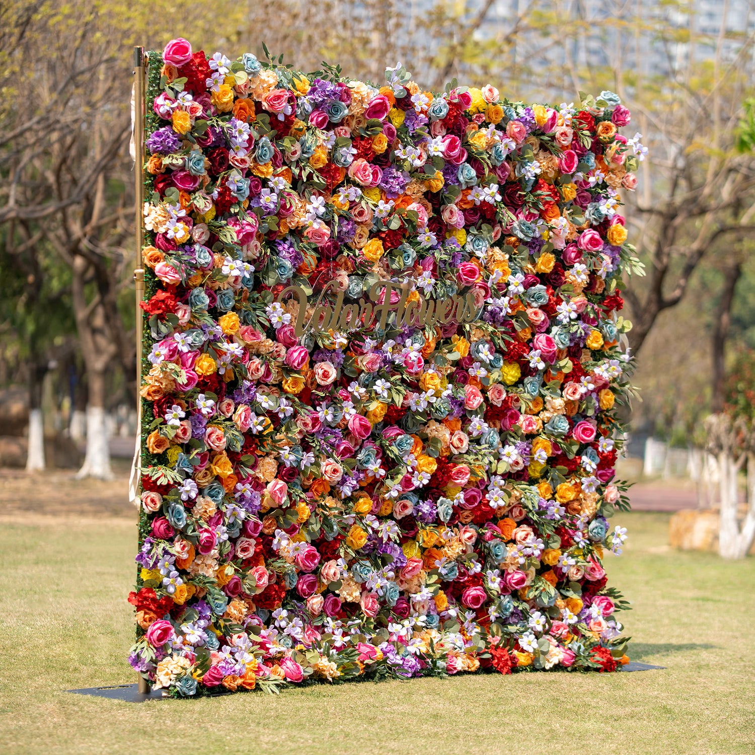 This image features a vibrant floral backdrop densely packed with multicolored roses in shades of red, orange, pink, purple, and yellow. The lush arrangement includes the words "Valar Flowers" in gold, set against an outdoor park - like background.