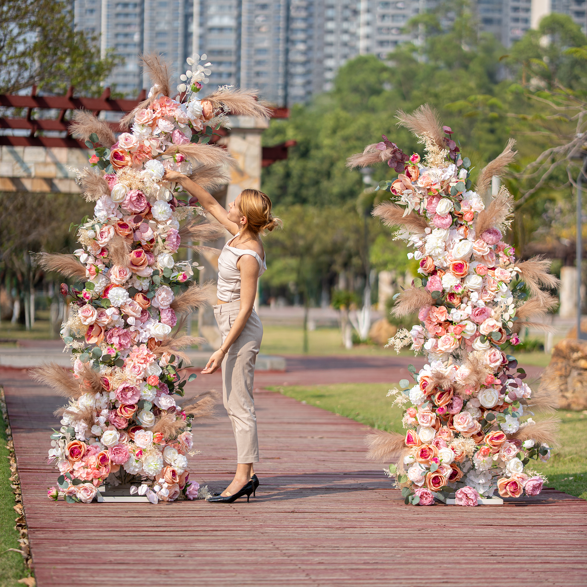 A woman in a sleeveless top and beige pants adjusts a floral archway. It features a mix of roses in pink, white, and peach hues, along with pampas grass. Set against an outdoor park - like backdrop, it&