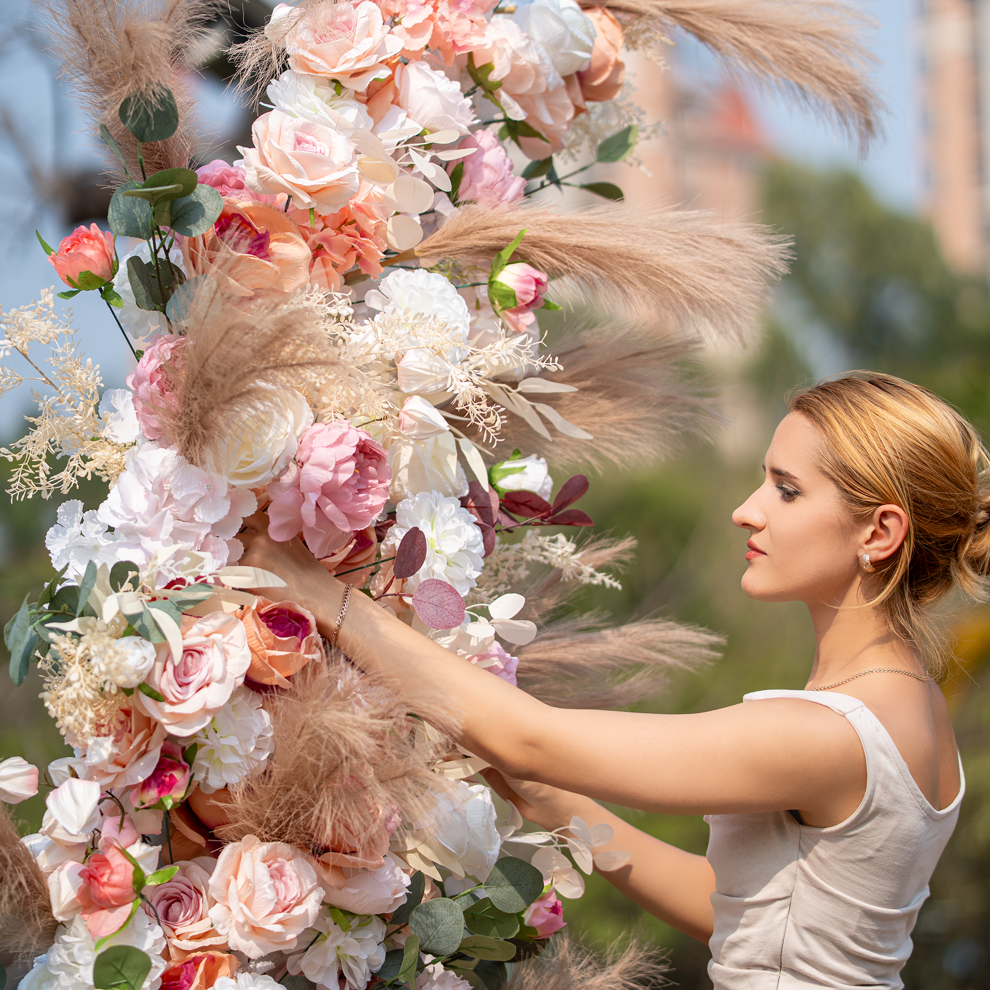 A woman arranges a lush floral installation outdoors. It combines roses, peonies, and pampas grass in soft pink, white, and beige hues. The backdrop features greenery and blurred buildings. Elegant and romantic, it&