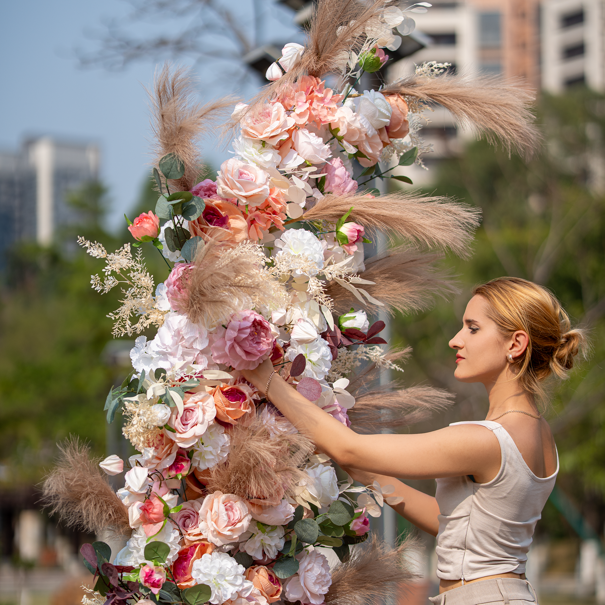 A woman in a sleeveless top and beige pants arranges a floral structure outdoors. It features roses, peonies, and pampas grass in soft pink, white, and beige tones. The backdrop includes greenery and buildings. Ideal for weddings or garden parties, it exudes a romantic, elegant style.