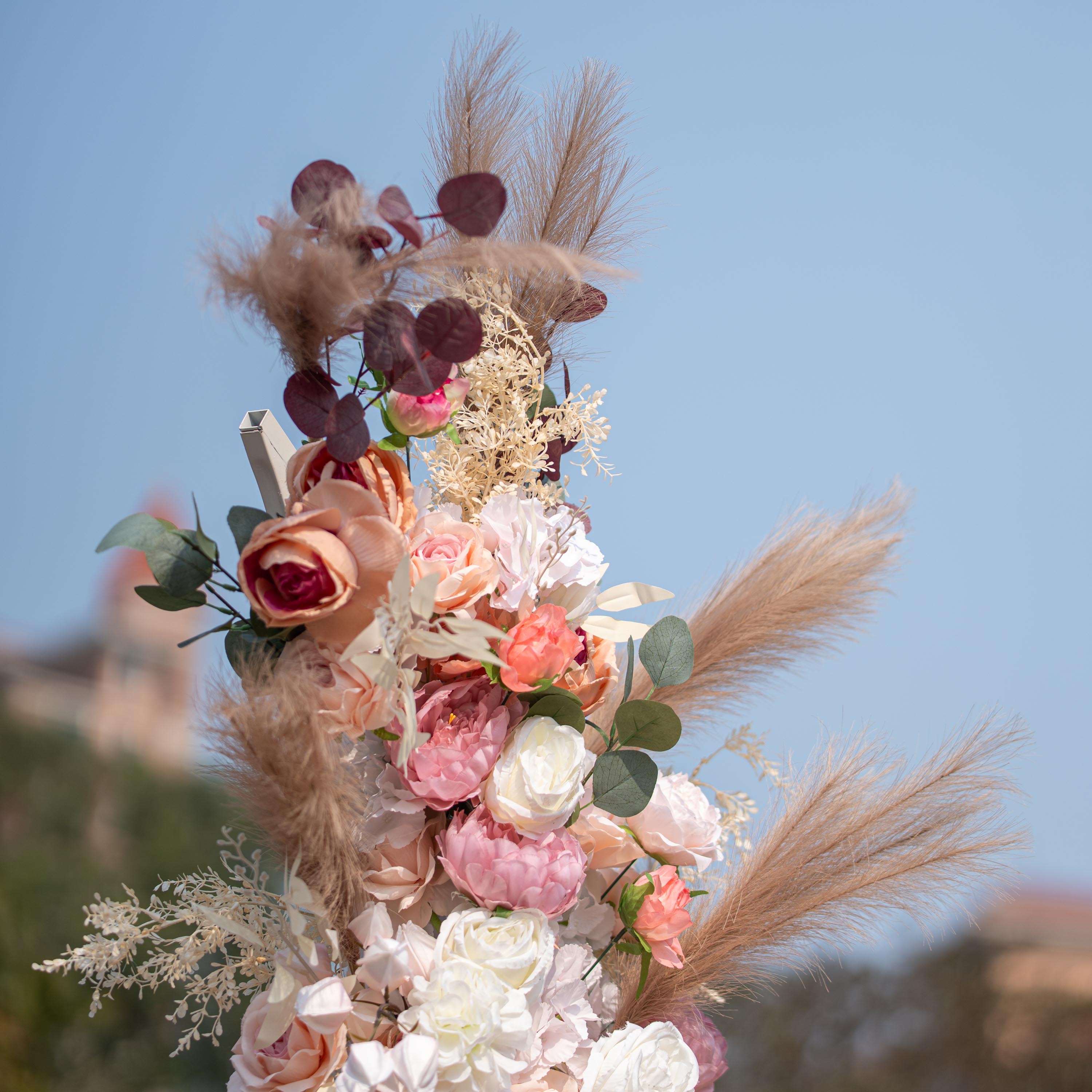 This photo shows a close - up of a floral arrangement with roses and peonies in soft pink, peach, and white tones. Pampas grass and eucalyptus leaves add texture. Set against a clear blue sky, it has a romantic, elegant style, perfect for weddings or upscale garden events.