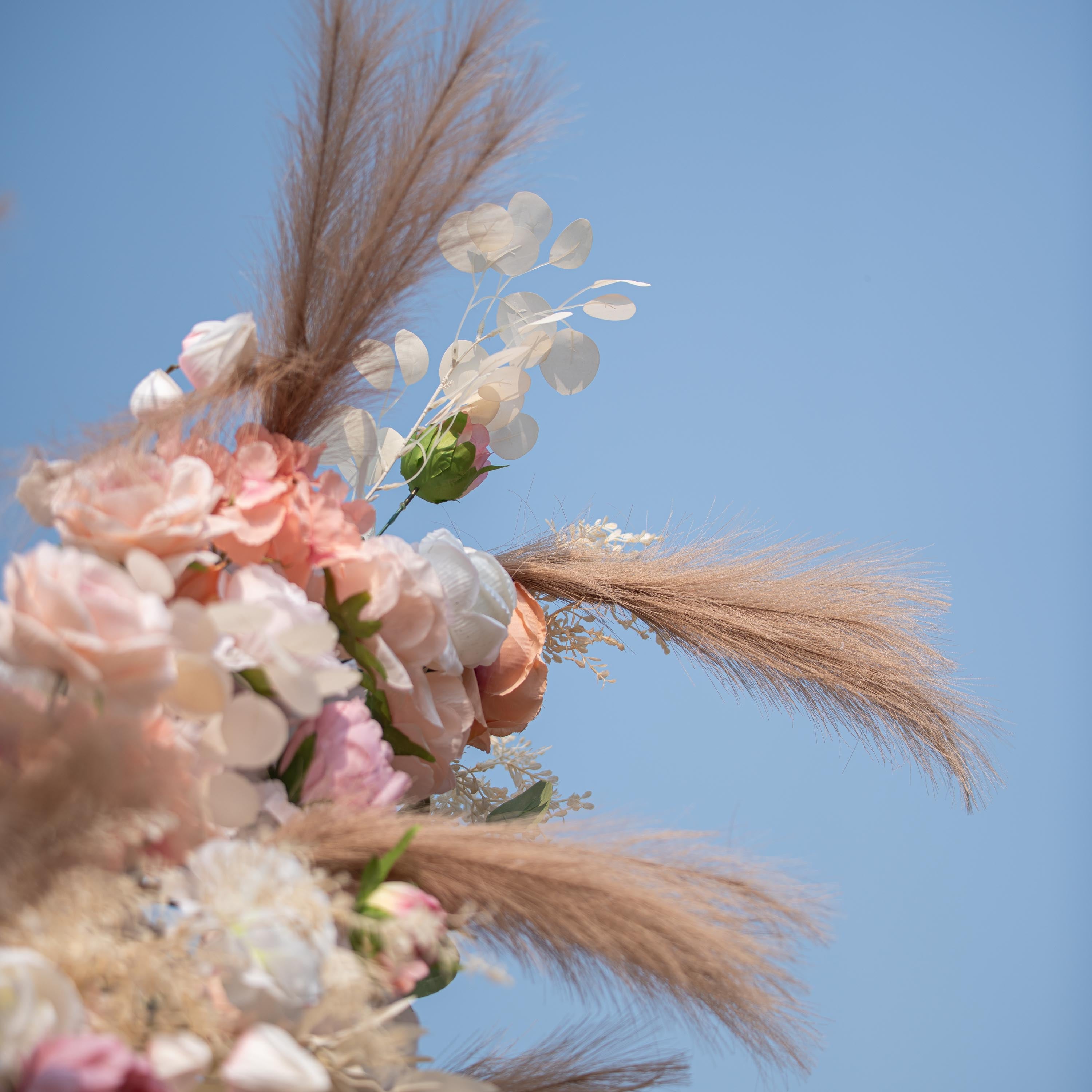 This photo captures a close - up of a floral arrangement featuring roses in soft pink and peach shades, along with white blooms. Pampas grass and eucalyptus leaves add texture. Set against a clear blue sky, it exudes a romantic, elegant style, perfect for weddings or garden parties.