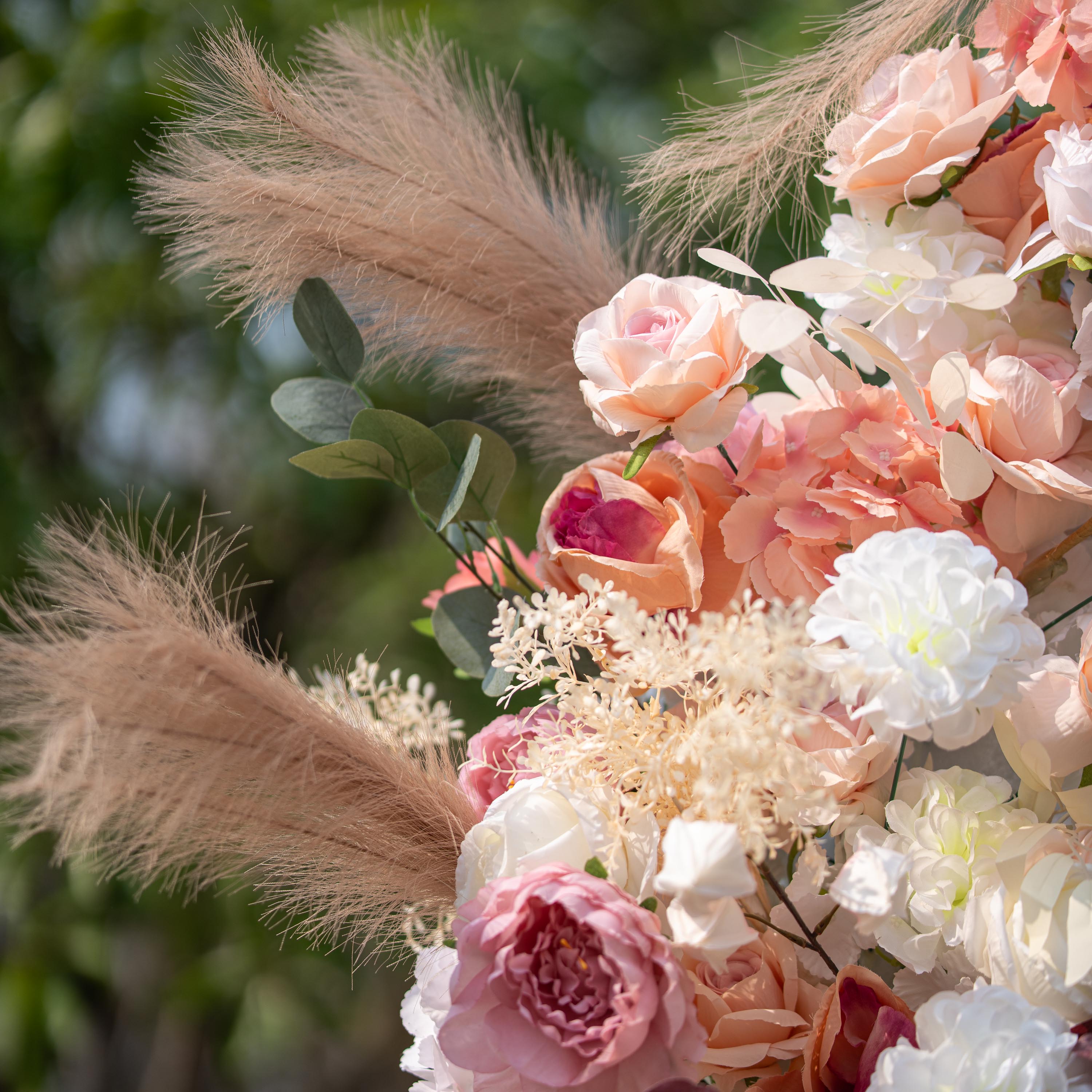 This close - up photo features a floral arrangement with roses in soft pink and peach tones, along with white blooms. Pampas grass and eucalyptus leaves add texture. Set against a blurred green outdoor backdrop, it has a romantic, elegant style, perfect for weddings or garden - themed events.