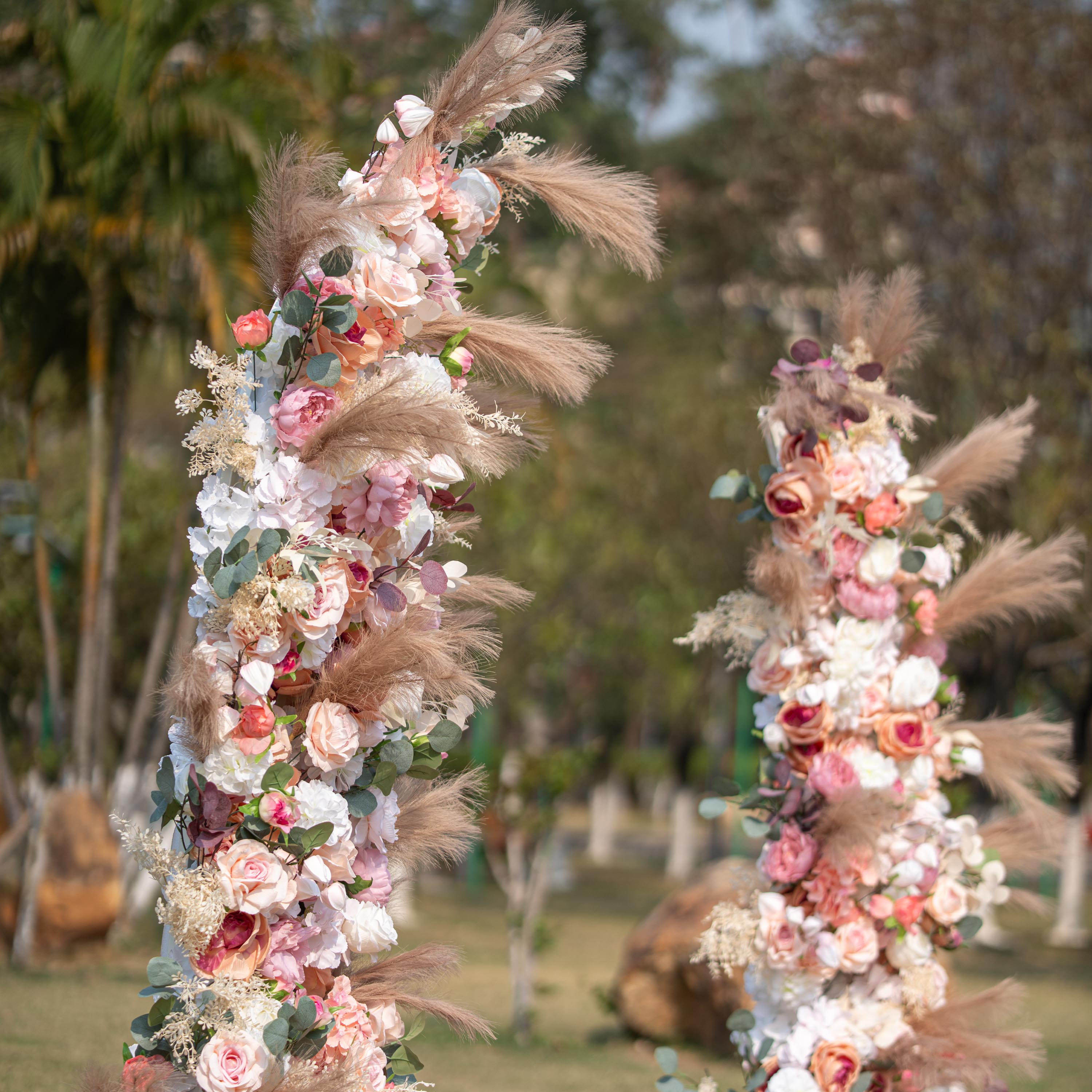 This photo showcases a floral arch adorned with roses and peonies in soft pink, peach, and white hues. Pampas grass and eucalyptus leaves add texture. Set outdoors with greenery in the background, it has a romantic, elegant style, perfect for weddings or garden parties.