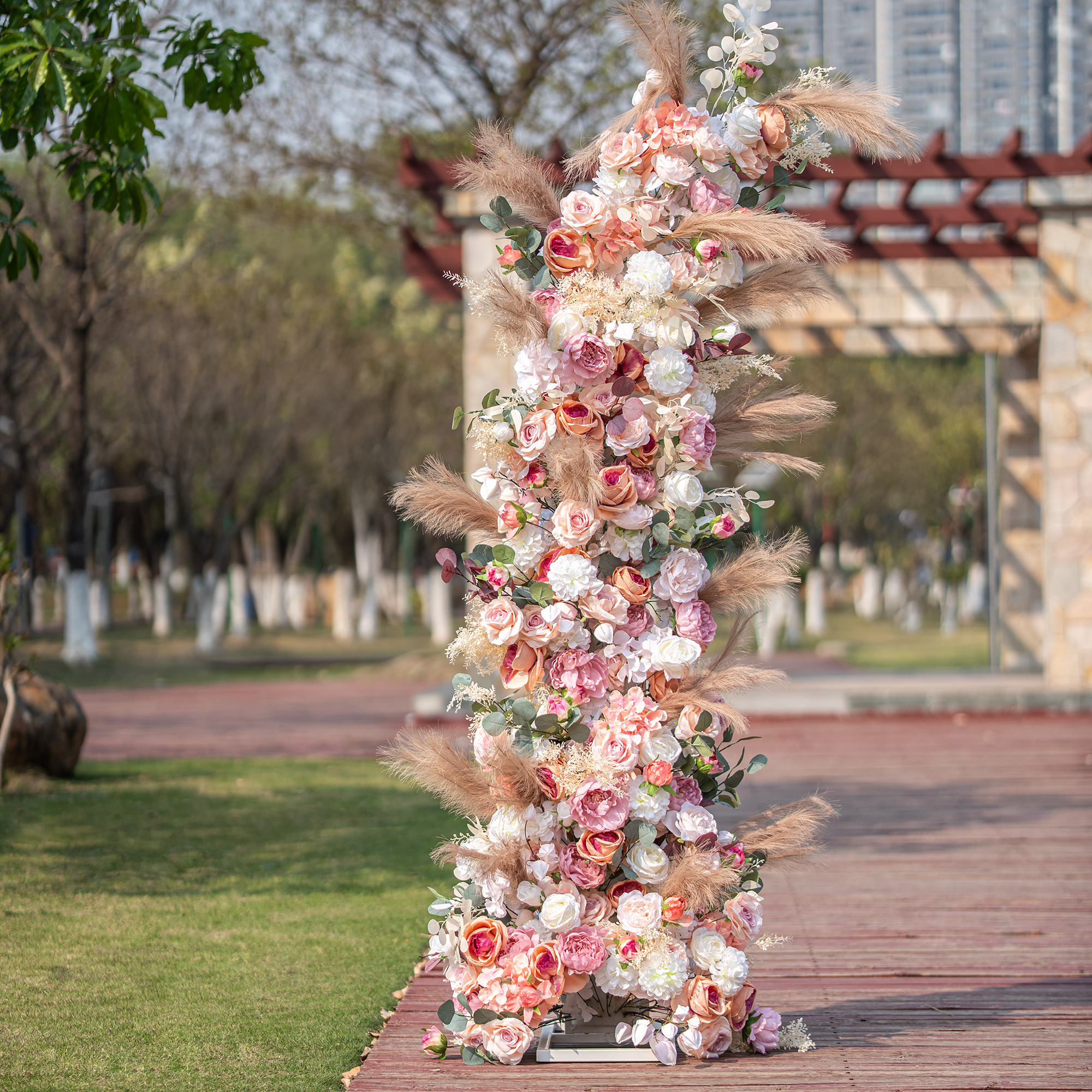 This photo features a tall floral arrangement, likely part of an arch, brimming with roses and peonies in soft pink, peach, and white shades. Pampas grass and eucalyptus leaves add texture. Set outdoors with greenery and a structure in the background, it has a romantic, elegant style, perfect for weddings.