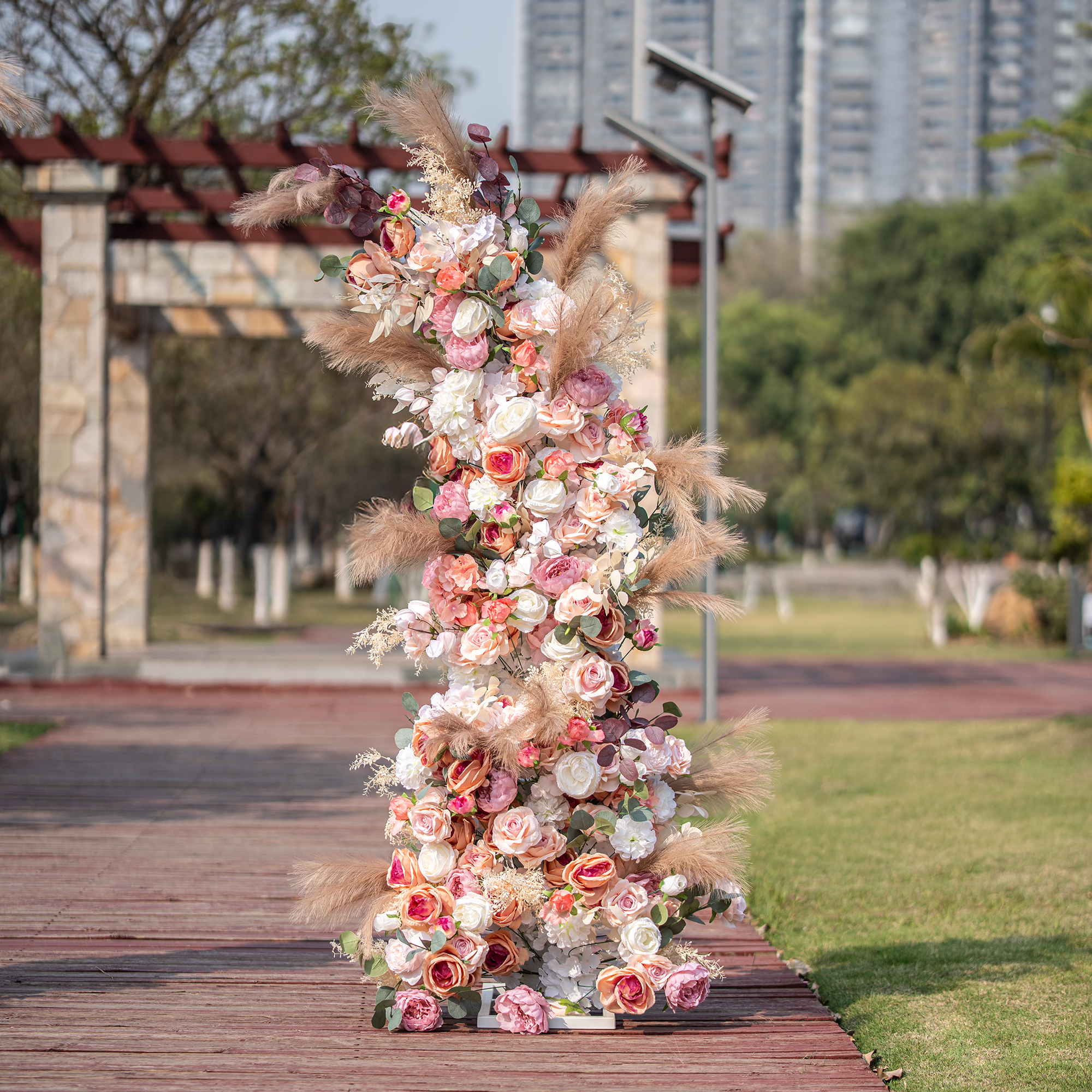 This photo showcases a tall, elaborate floral arrangement, likely part of an arch, bursting with roses and peonies in soft pink, peach, and white tones. Pampas grass and eucalyptus leaves add texture. Set outdoors with greenery and city buildings in the background, it has a romantic, elegant style, perfect for weddings or upscale garden events.