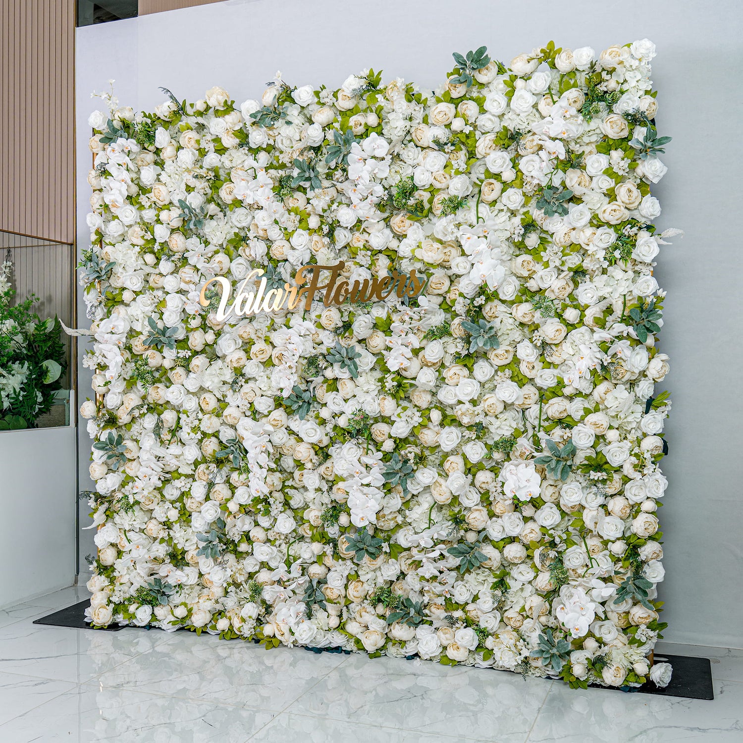This backdrop is a lush, dense arrangement of white artificial flowers interspersed with green foliage. The elegant display creates a soft, romantic atmosphere, with the golden "Valar Flowers" sign adding a touch of luxury, perfect for weddings or upscale events.