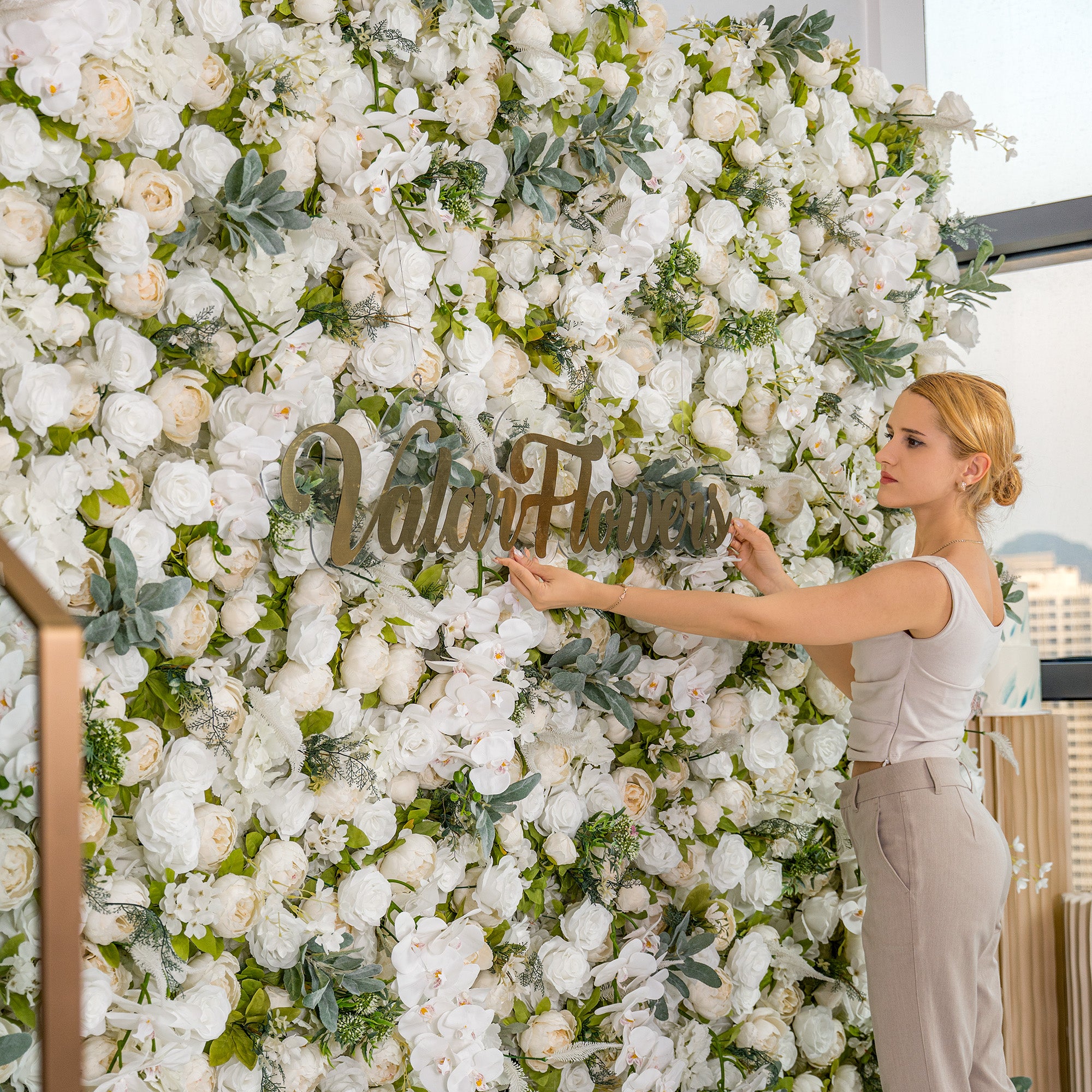 A woman adjusts a “Valar Flowers” sign on a lush floral backdrop. The backdrop is filled with white roses, peonies, and orchids interspersed with green foliage, creating an elegant and romantic setting.
