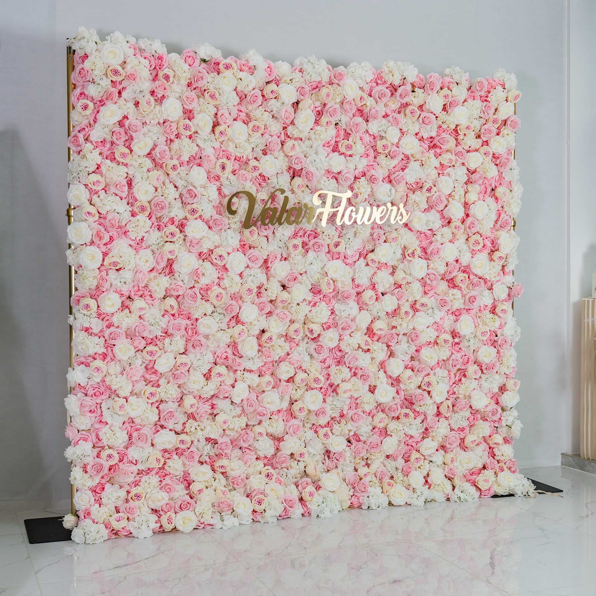 This photo features a lush floral backdrop composed predominantly of pink and white roses. The dense arrangement creates a soft, dreamy effect with its pastel color palette. The elegant gold text "Valar Flowers" is prominently displayed in the center. Ideal for weddings, bridal showers, or other romantic events, it exudes a romantic and sophisticated style.