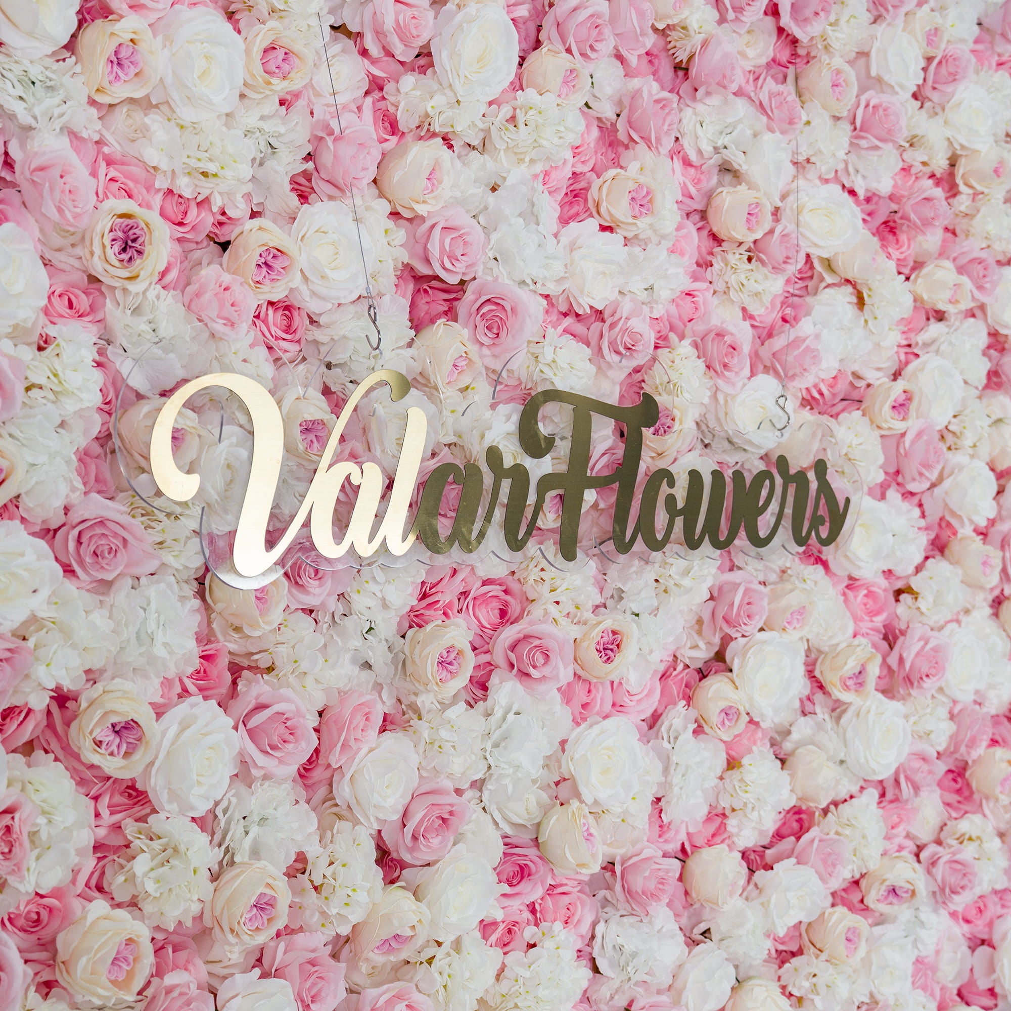 This photo showcases a lush floral backdrop densely packed with pink and white roses, along with white hydrangeas. The soft pastel color palette creates a romantic and elegant atmosphere. The words "Valar Flowers" in elegant gold lettering stand out prominently. Ideal for weddings, bridal showers, or other romantic events, it exudes a dreamy and sophisticated style.