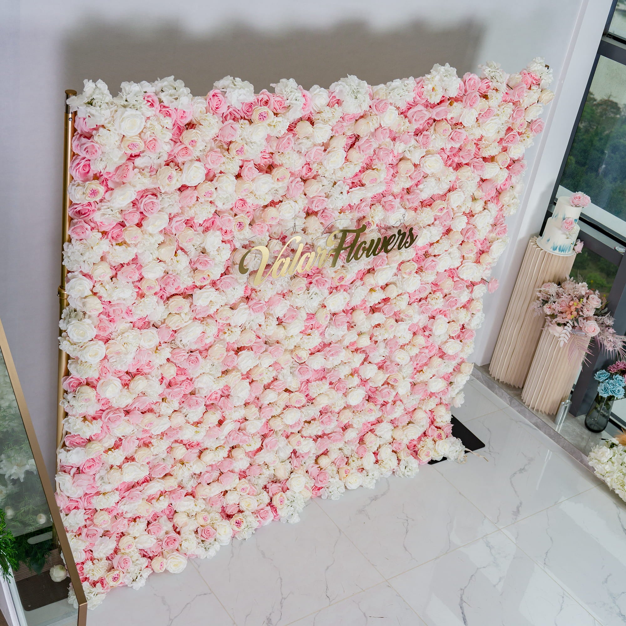 This photo features a lush floral backdrop densely covered with pink and white roses, creating a soft and romantic aesthetic. The pastel color palette evokes a dreamy atmosphere. The elegant gold text "Valar Flowers" stands out prominently. Ideal for weddings, bridal showers, or other romantic events, it exudes a sophisticated and tender style.