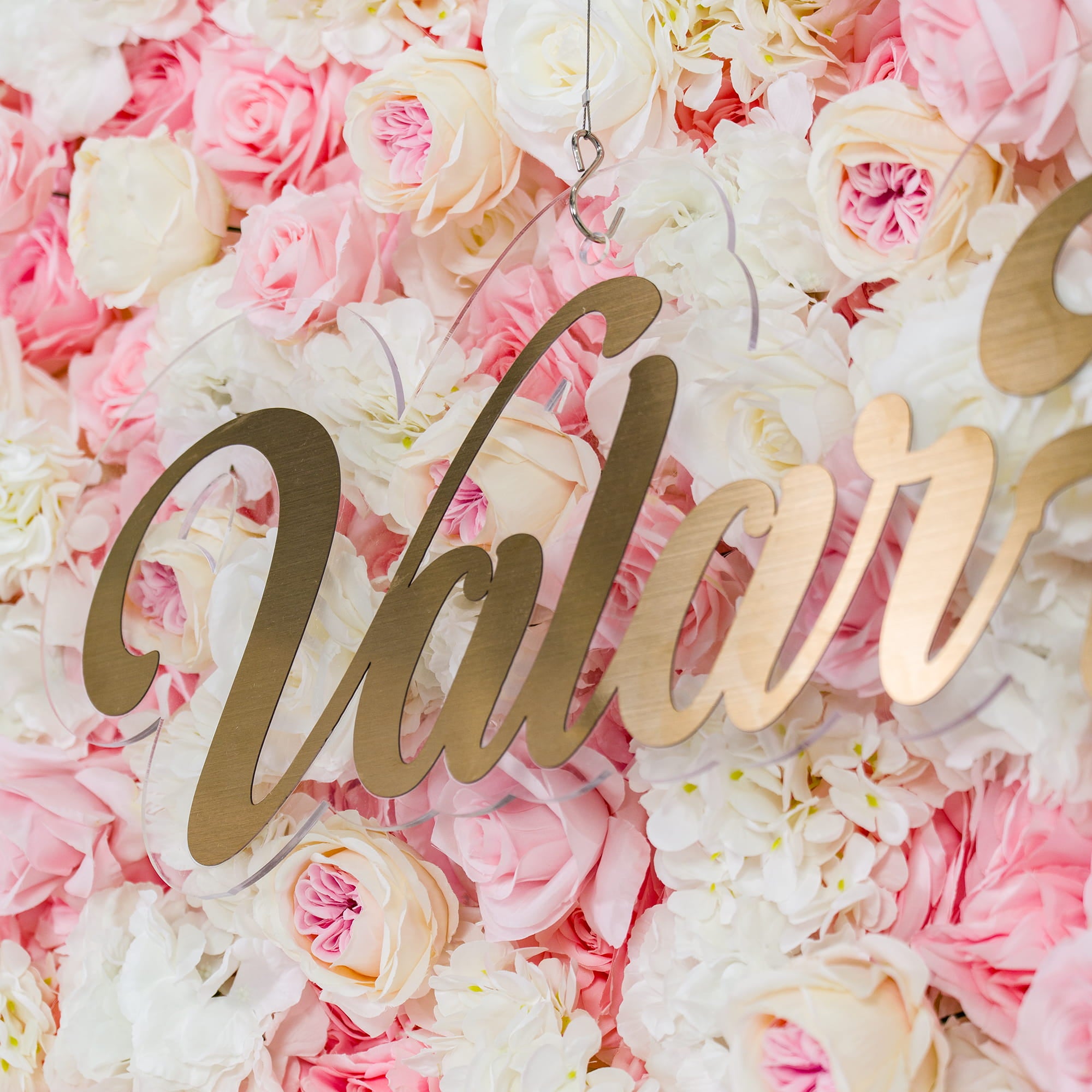 This photo showcases a lush floral backdrop with a dense arrangement of pink and white roses. The soft pastel color palette creates a romantic and elegant atmosphere. The word "Valar" in elegant gold lettering stands out prominently. Ideal for weddings or bridal showers, it exudes a dreamy and sophisticated style.