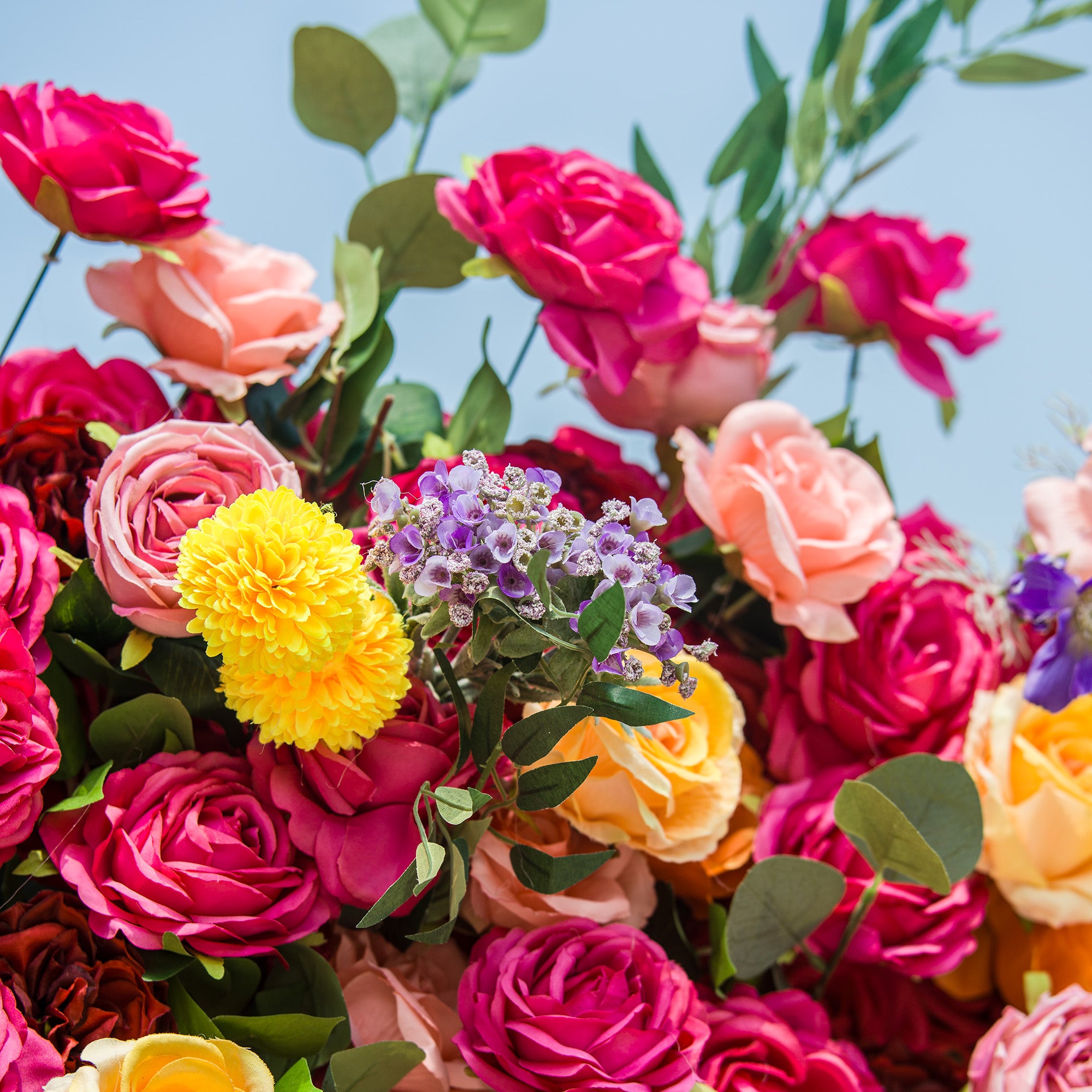 A vibrant floral arrangement bursts with color against a bright sky. Deep pink roses dominate, interspersed with soft peach and yellow blooms. A cluster of purple flowers and fluffy yellow blossoms add contrast, while eucalyptus leaves provide fresh greenery. The dense, varied blooms create a lively, eye-catching display, perfect for celebrations or decorative focal points.
