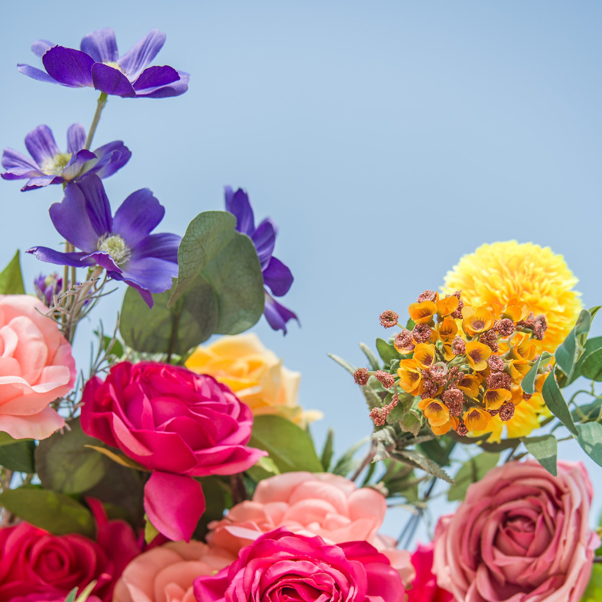 A vibrant floral arrangement bursts with color against a bright blue sky. Pink and magenta roses take center stage, surrounded by soft peach - toned blooms. Yellow flowers, purple blossoms, and green foliage add texture and contrast. The mix of large, showy roses and delicate smaller flowers creates a lively, eye - catching display, perfect for adding a touch of natural beauty to any setting.
