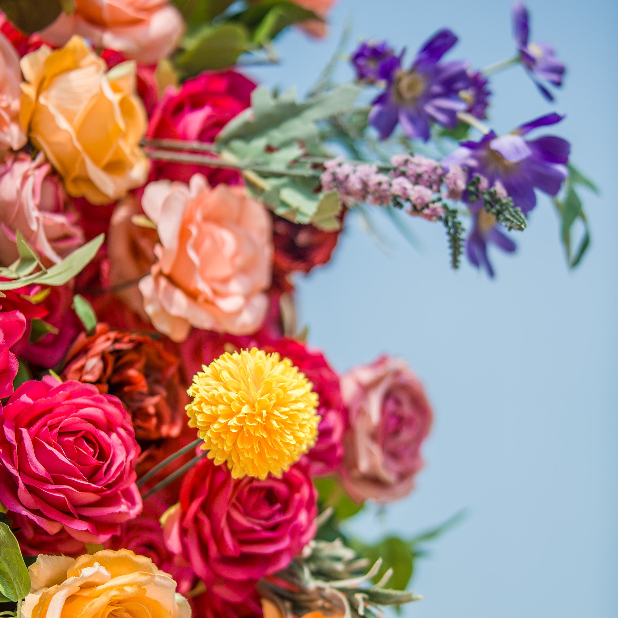 A vibrant floral arrangement bursts with color against a bright blue sky. Deep pink roses, soft peach blooms, and sunny yellow flowers create a lively base. Purple blossoms and delicate lavender - hued sprigs add pops of cool tones, while green foliage weaves through, enhancing the natural, eye - catching display—perfect for adding a romantic, cheerful touch to any space.
