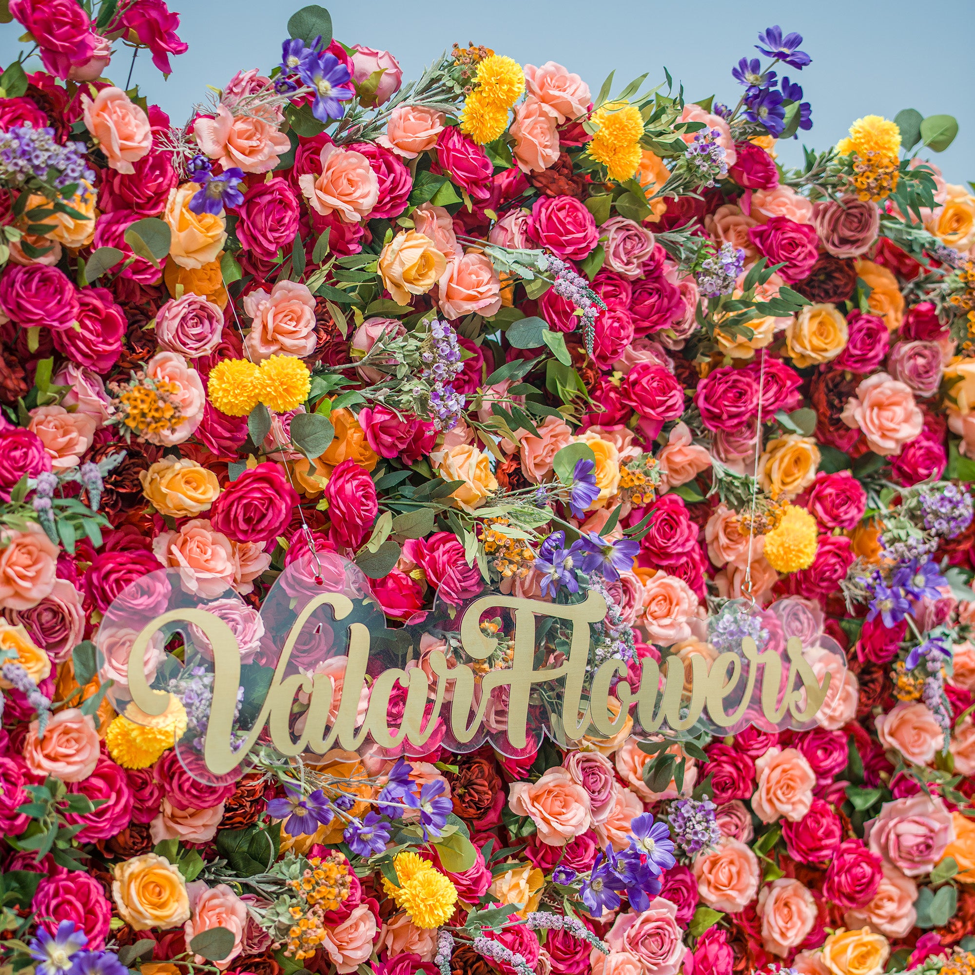 A vibrant floral wall by ValarFlowers, overflowing with colorful blooms! Rich red roses, soft peach roses, sunny yellow flowers, and purple blossoms create a lush backdrop. The gold “ValarFlowers” logo on clear acrylic stands out, blending natural beauty with brand charm. Perfect for events, this eye - catching display offers a picturesque, Instagram - worthy setting.
