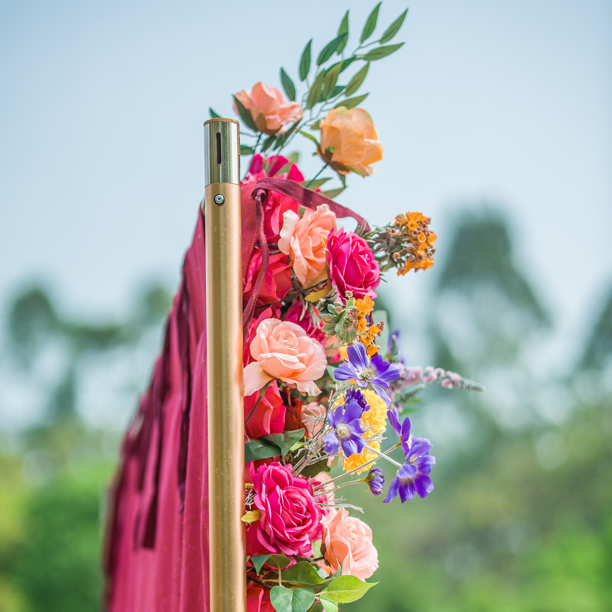 A close - up of a decorative floral arrangement on a gold pole. It features a mix of pink, peach, and yellow roses, along with purple flowers and orange blossoms. The burgundy fabric and green foliage add texture, set against a soft, blurred natural background. Perfect for events, it combines elegance and color in a charming display.