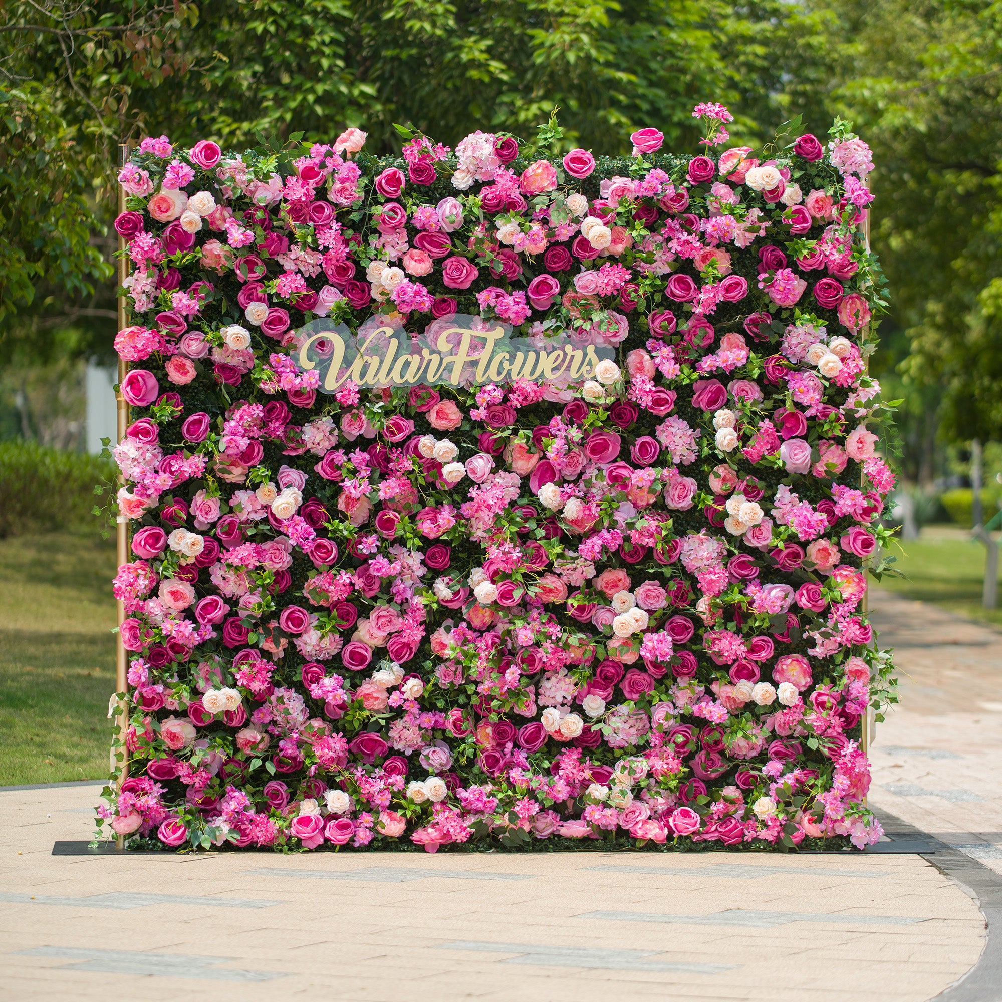 This photo features an 8X8FT floral backdrop with a golden stand, densely adorned with an array of pink and white roses and other pink flowers amidst lush green foliage. The text “ValarFlowers” in elegant golden lettering stands out prominently. Set outdoors with greenery in the background, it exudes a lively, romantic charm, perfect for events like garden weddings.
