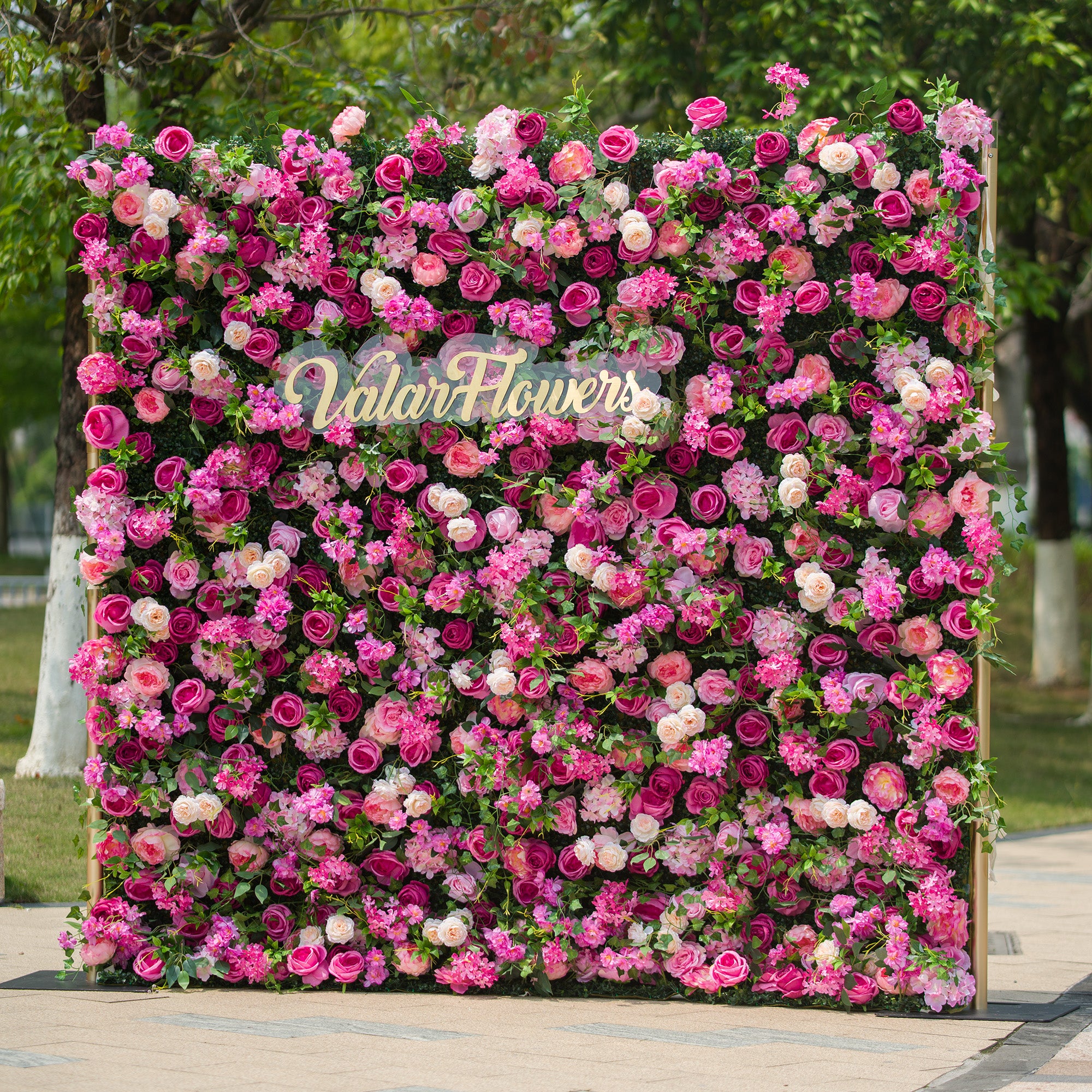 This photo showcases a large, vibrant floral backdrop brimming with an array of pink and white roses, along with other assorted flowers, all nestled among lush green foliage. The text "ValarFlowers" is prominently displayed in elegant golden lettering in the center. Set outdoors with greenery in the background, it exudes a lively and romantic charm, perfect for events like garden weddings or spring festivals.