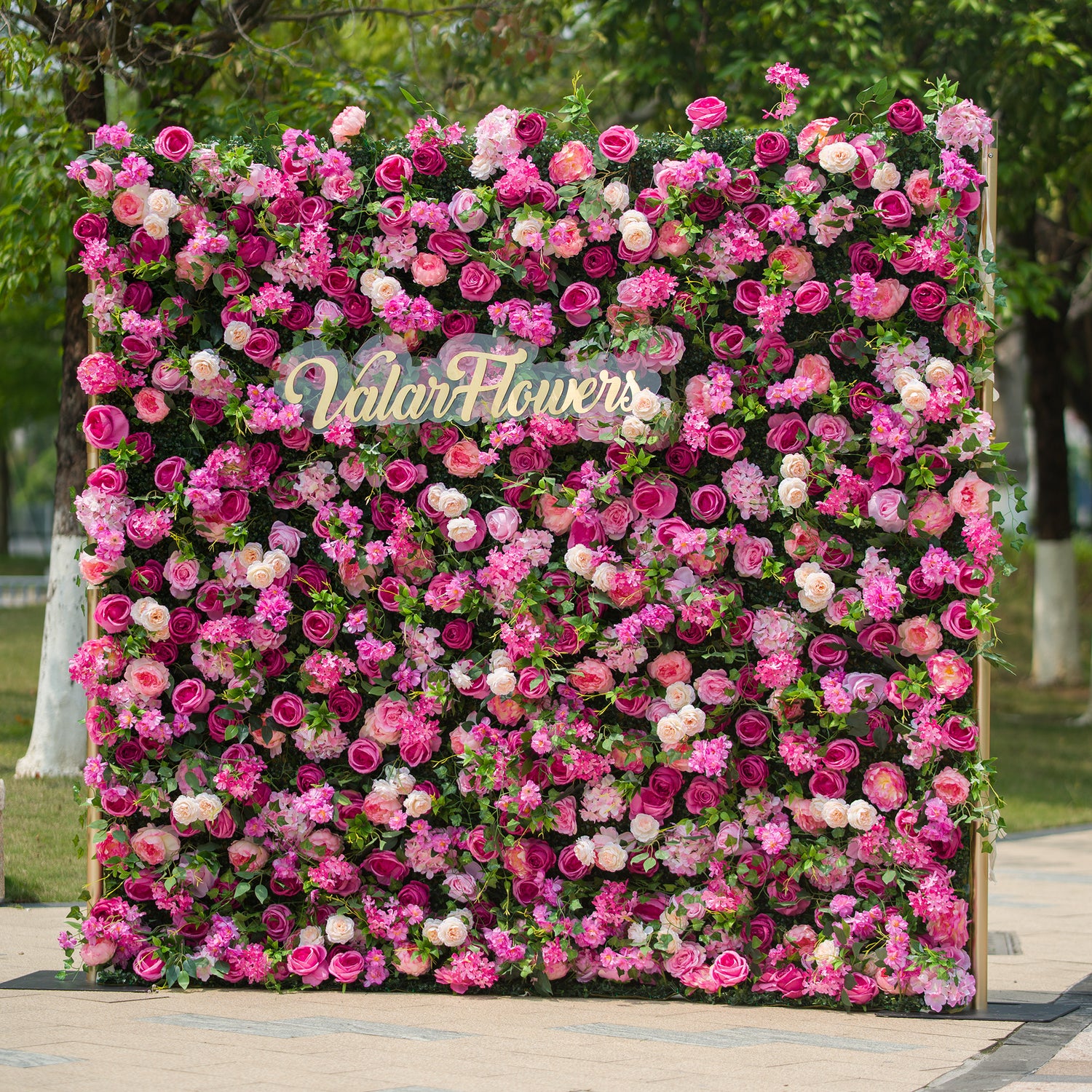 This photo showcases a large, vibrant floral backdrop brimming with an array of pink and white roses, along with other assorted flowers, all nestled among lush green foliage. The text "ValarFlowers" is prominently displayed in elegant golden lettering in the center. Set outdoors with greenery in the background, it exudes a lively and romantic charm, perfect for events like garden weddings or spring festivals.