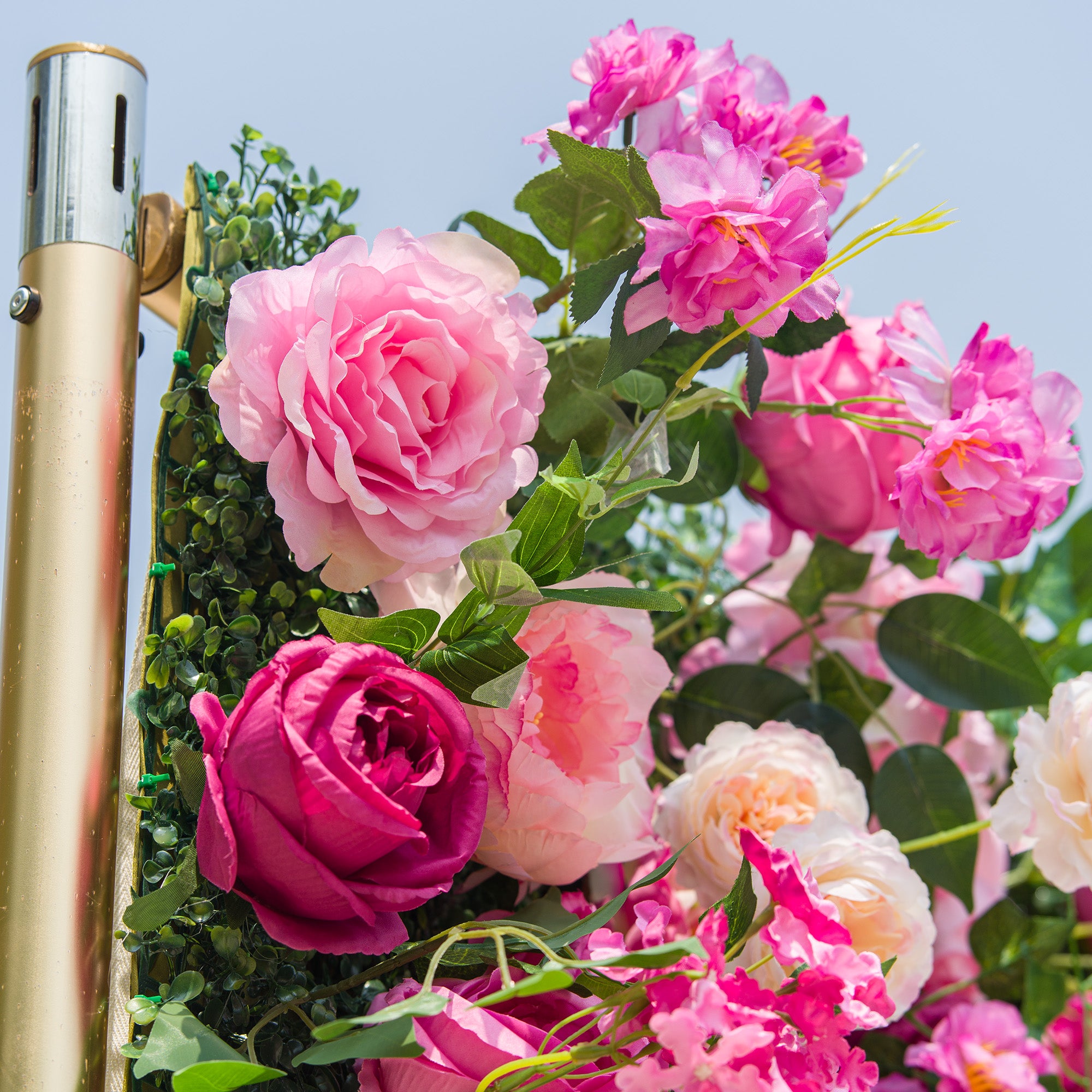 This photo showcases a vibrant floral arrangement featuring an array of pink roses in various shades, from soft pastels to deep magenta, along with some white roses. The flowers are interspersed with lush green foliage, creating a striking contrast. Set against a clear sky, with a metallic pole partially visible, the arrangement exudes a lively and romantic charm, perfect for outdoor events like garden weddings or summer parties.