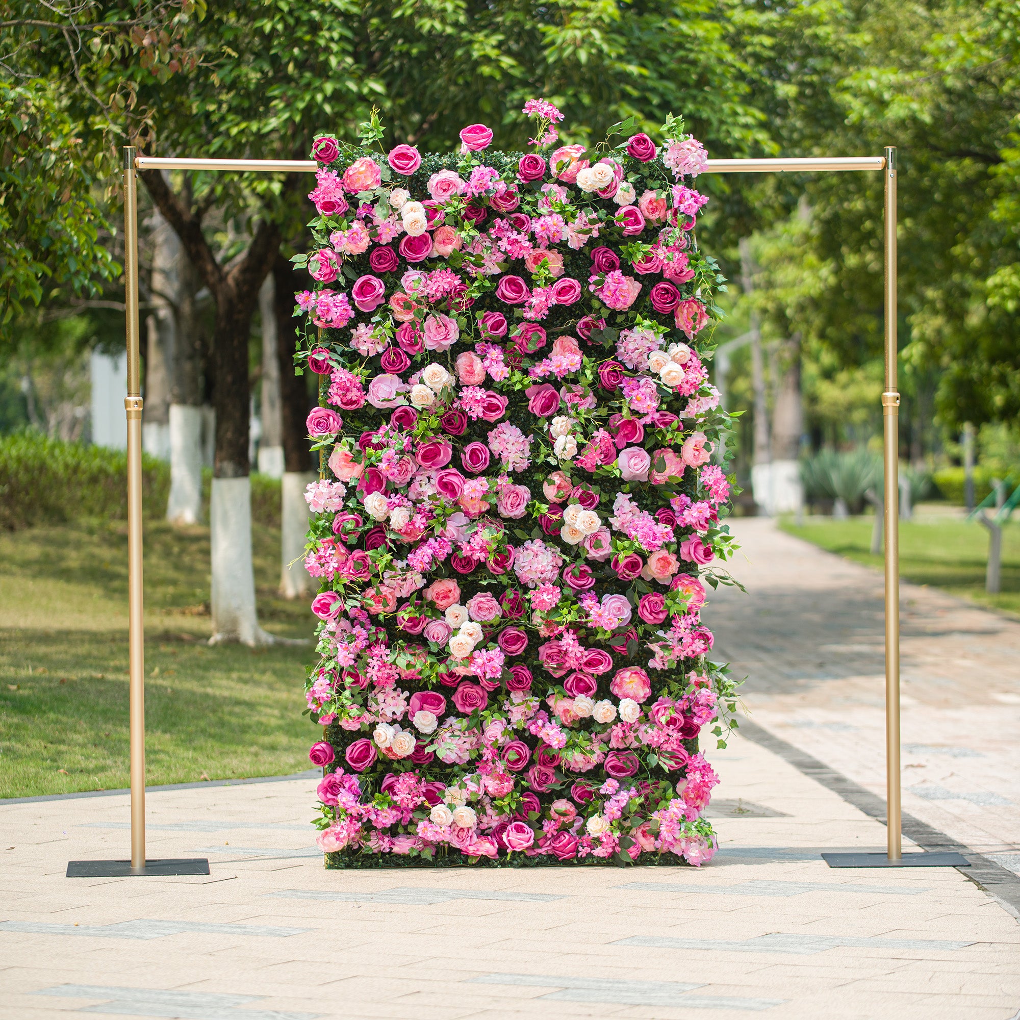 This photo showcases a 4X8ft floral backdrop mounted on a golden stand. It is densely covered with a vibrant mix of pink and white roses and other pink flowers, interspersed with lush green foliage. Set outdoors on a paved path with greenery in the background, it exudes a lively and romantic charm, ideal for events like weddings or garden parties.