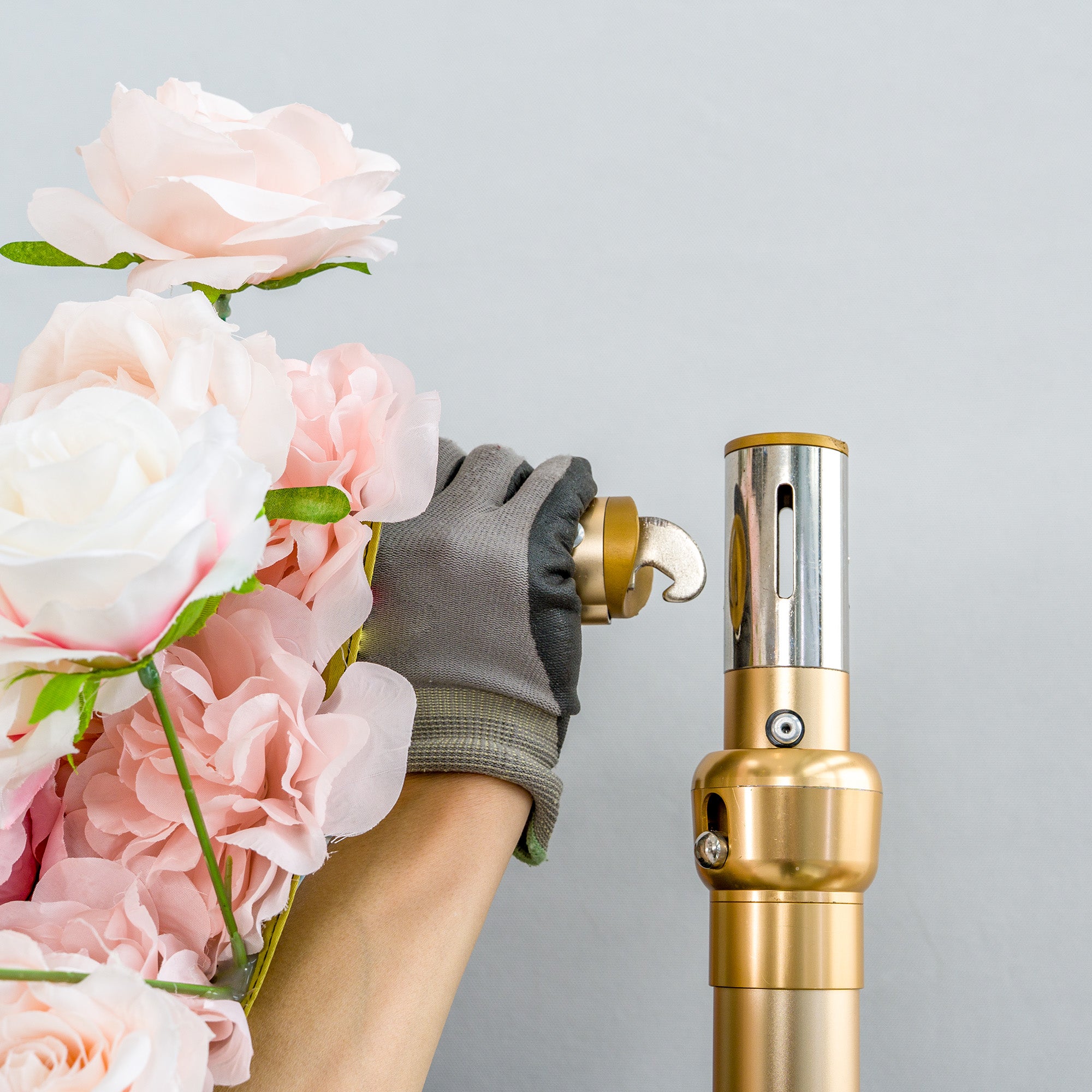 This photo captures a close - up scene of floral arrangement work. A gloved hand, wearing a grey glove, is seen handling pink and white artificial roses. The hand is positioned next to a golden - colored metal pole, which likely serves as part of the structure for supporting floral displays. The background is a plain, light grey color, highlighting the focus on the hand, flowers, and the metallic component, suggesting a behind - the - scenes look at setting up decorative floral installations.