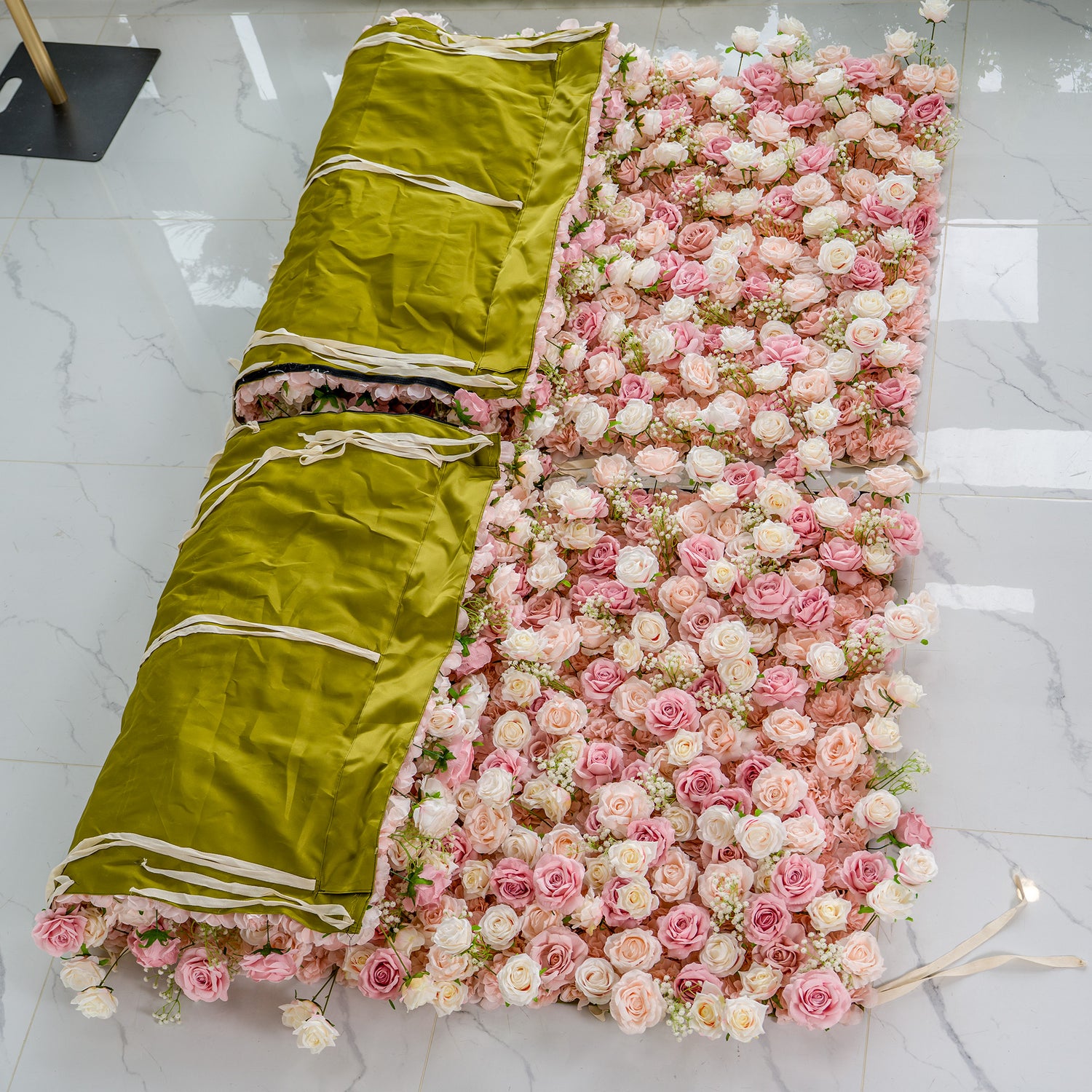 This photo shows a floral backdrop made of pink and white roses, partially rolled up. The rolled sections are covered with a green fabric secured by white straps. The backdrop is laid out on a white marble - tiled floor. This setup likely indicates storage or transport of the floral arrangement, which is typically used for events like weddings to create a romantic and elegant atmosphere.