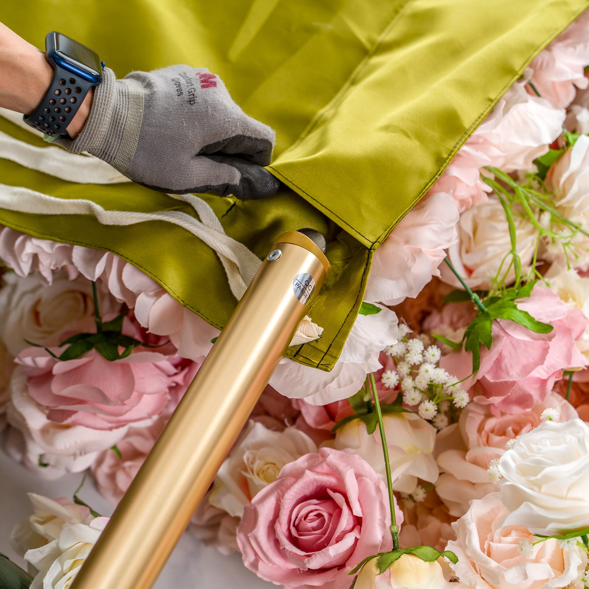 This photo captures a close - up scene of floral backdrop handling. A gloved hand, wearing a smartwatch, is seen adjusting a green fabric cover over a lush arrangement of pink and white roses. The hand is positioned next to a golden - colored metal pole, which is partially covered by the fabric. The focus is on the process of securing or uncovering the floral display, highlighting the care taken in setting up or storing the decorative piece.
