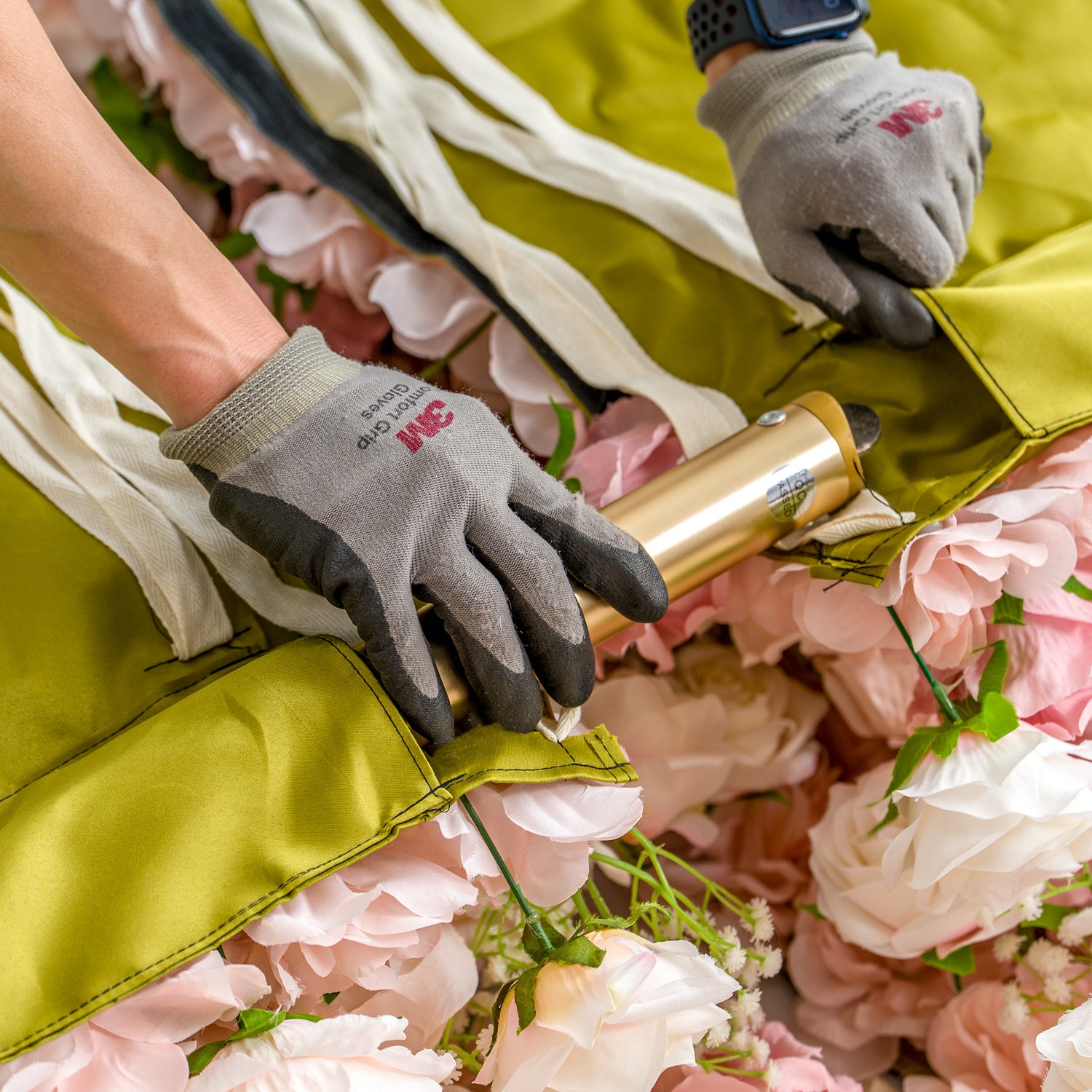 This photo shows a close - up of two gloved hands working on a floral arrangement. The gloves are branded "Safety Grip Gloves." The hands are adjusting a golden metal pole that is partially covered by a green fabric. Surrounding the pole are numerous pink and white roses, creating a lush, decorative backdrop. The scene captures the meticulous process of setting up or securing the floral display.