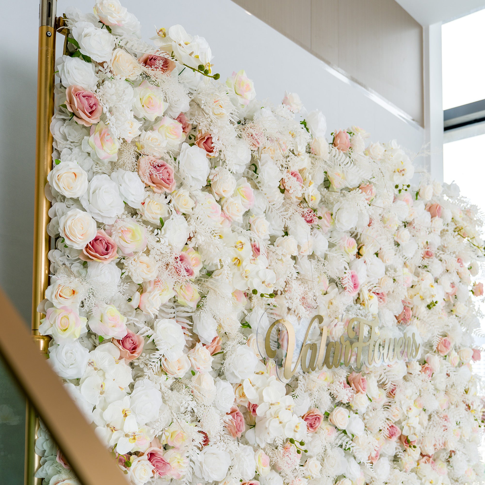 This photo showcases a lush floral backdrop densely covered with white roses, interspersed with delicate pink roses and some white foliage like ferns. The soft color palette of white and light pink creates an elegant and serene atmosphere. The words "Valar Flowers" in elegant gold lettering stand out. Ideal for weddings or upscale events, it exudes a refined, dreamy style.