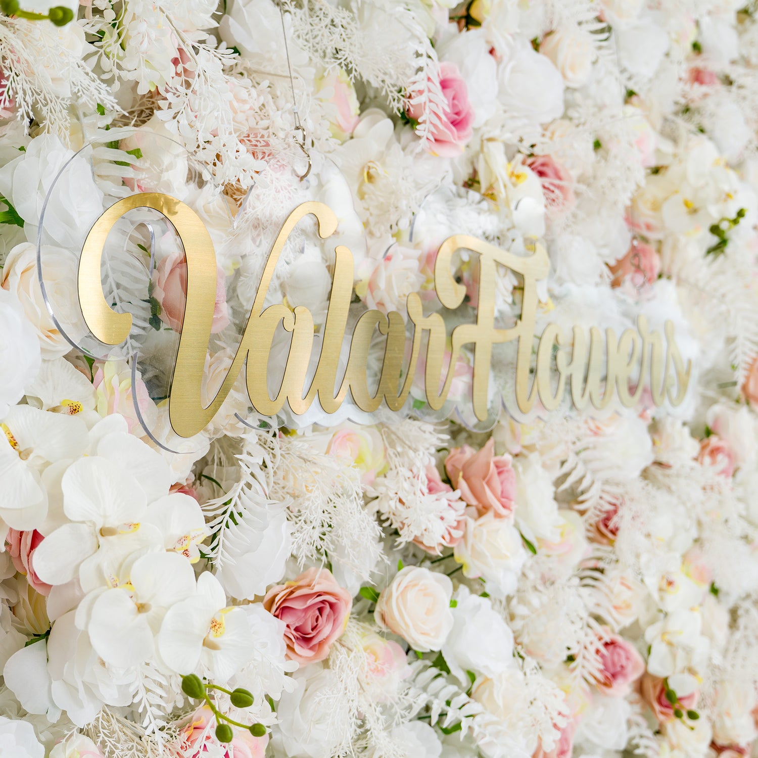 This photo showcases a lush floral backdrop filled with white roses, pink - tinged roses, and white orchids, complemented by delicate white foliage. The soft pastel color palette of white and light pink creates an elegant and romantic atmosphere. The words "Valar Flowers" in elegant gold lettering stand out prominently. Ideal for weddings or upscale events, it exudes a refined, dreamy style.