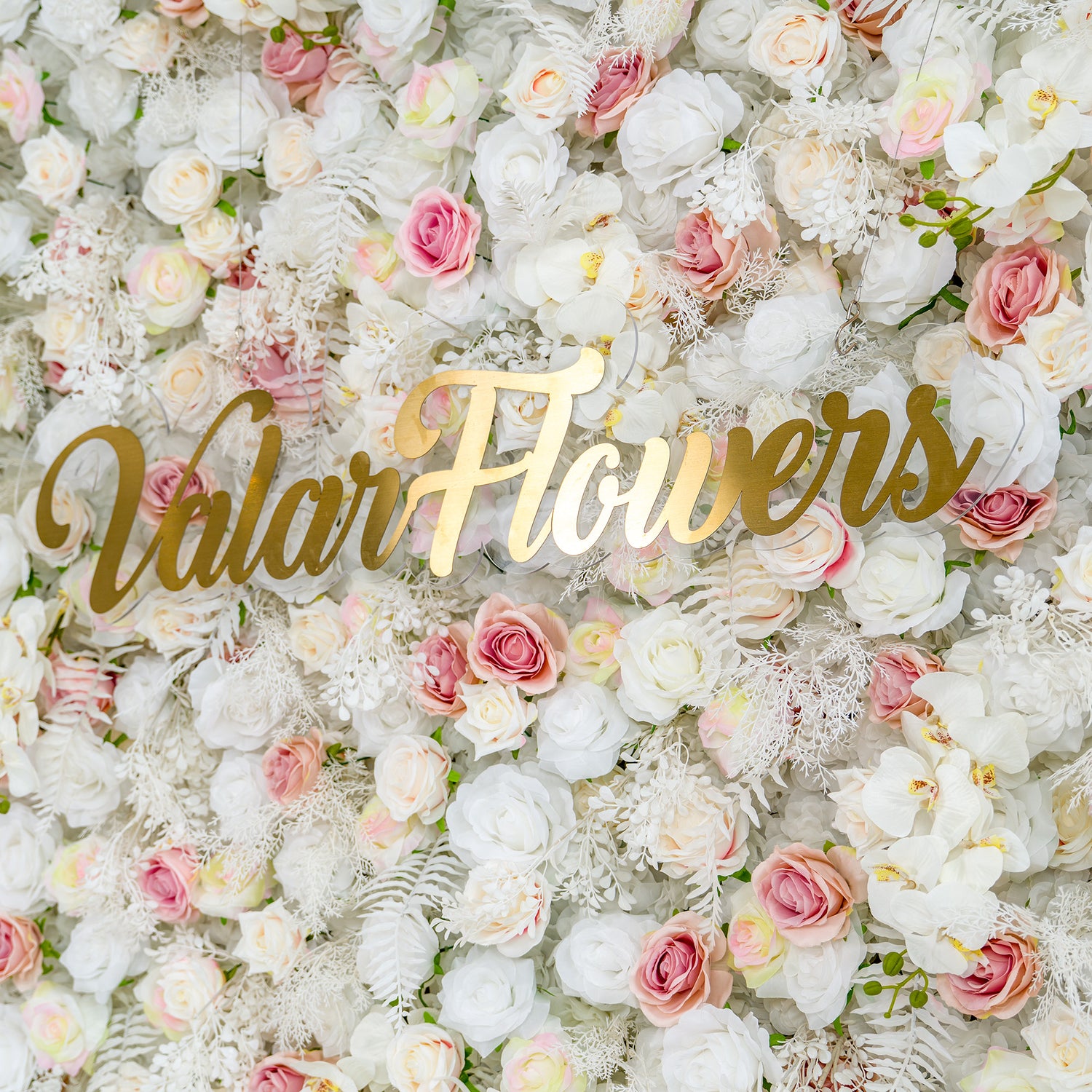 This photo showcases a lush floral backdrop with white roses, pink roses, and white orchids, complemented by white foliage. The soft color palette of white, light pink, and hints of pastel yellow creates an elegant and romantic atmosphere. The golden text "Valar Flowers" stands out prominently. Ideal for weddings or upscale events, it exudes a refined, dreamy style.