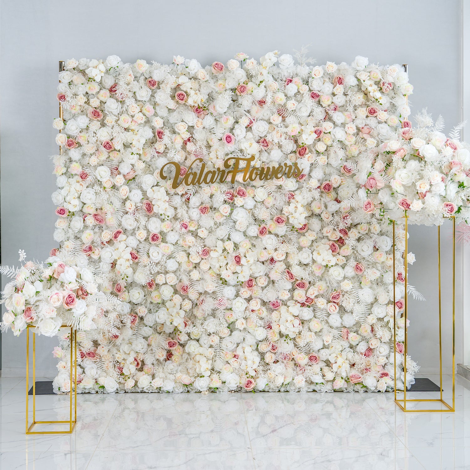 This photo features a large floral backdrop densely covered with white roses, interspersed with pink roses and white foliage. The soft color palette of white and light pink creates an elegant and serene atmosphere. The words "Valar Flowers" in gold lettering stand out prominently. Ideal for weddings or upscale events, it exudes a refined, dreamy style.